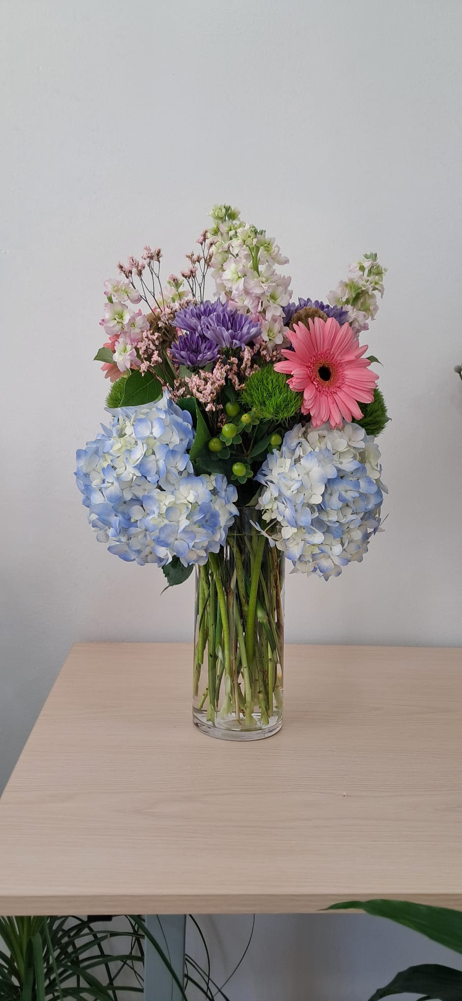 Mixed bouquet with blue hydrangeas, pink gerbera daisy, and purple blooms in a clear vase