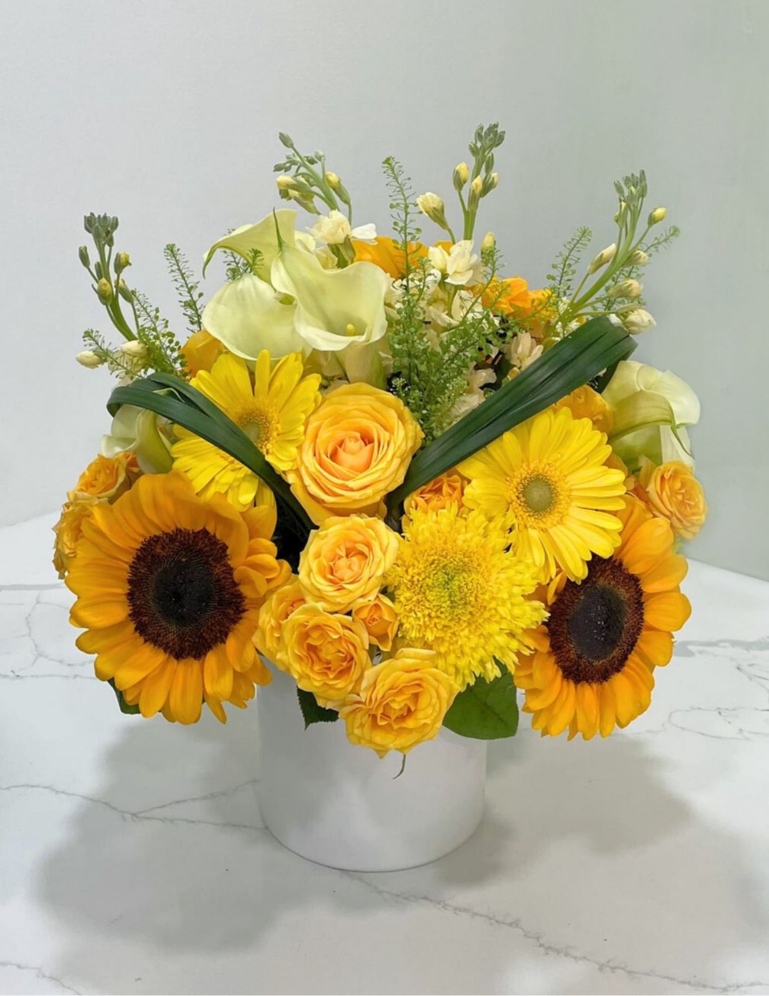 Finest Flowers - This beautiful arrangement has sun flowers, calla lilies, spray roses, roses, stock, lily grass, gerbera daisies, and mums in a white vase. 
