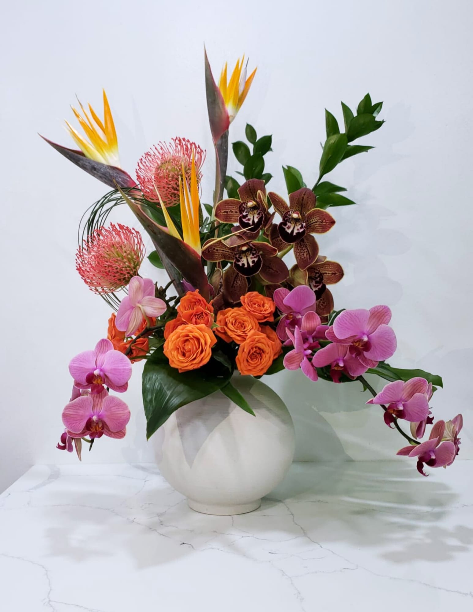 Tropical arrangement with orchids, orange roses, and bird-of-paradise in a white vase