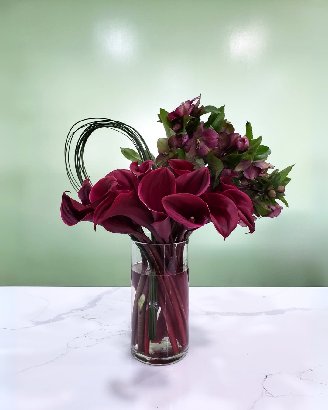 Deep burgundy calla lilies and purple flowers in a clear glass vase