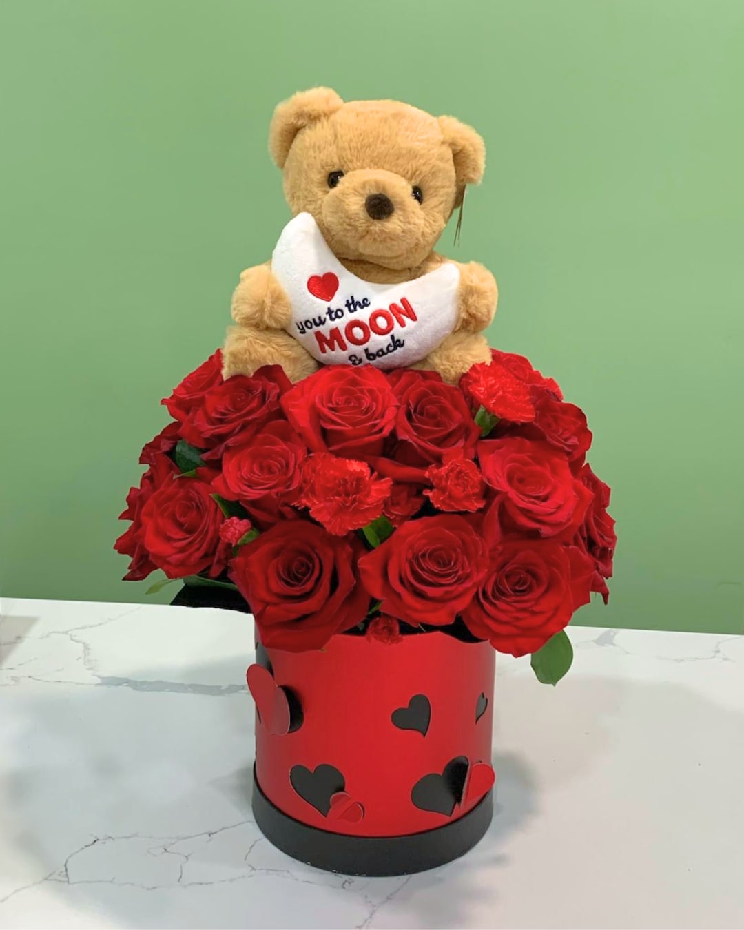 Teddy bear holding a heart pillow above red roses in a heart-decorated box