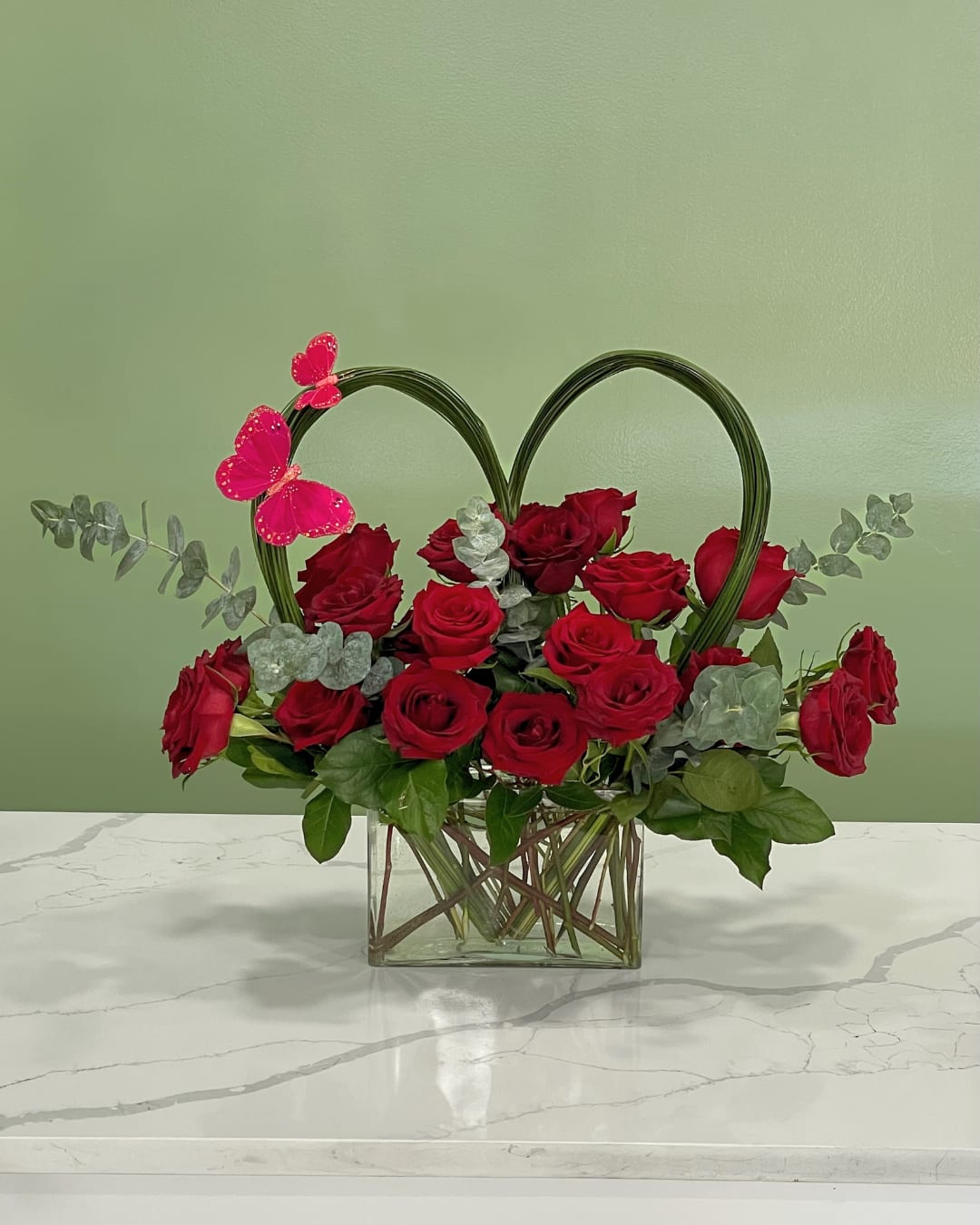 Red roses arranged in a glass vase with a heart-shaped frame and pink butterfly accents.