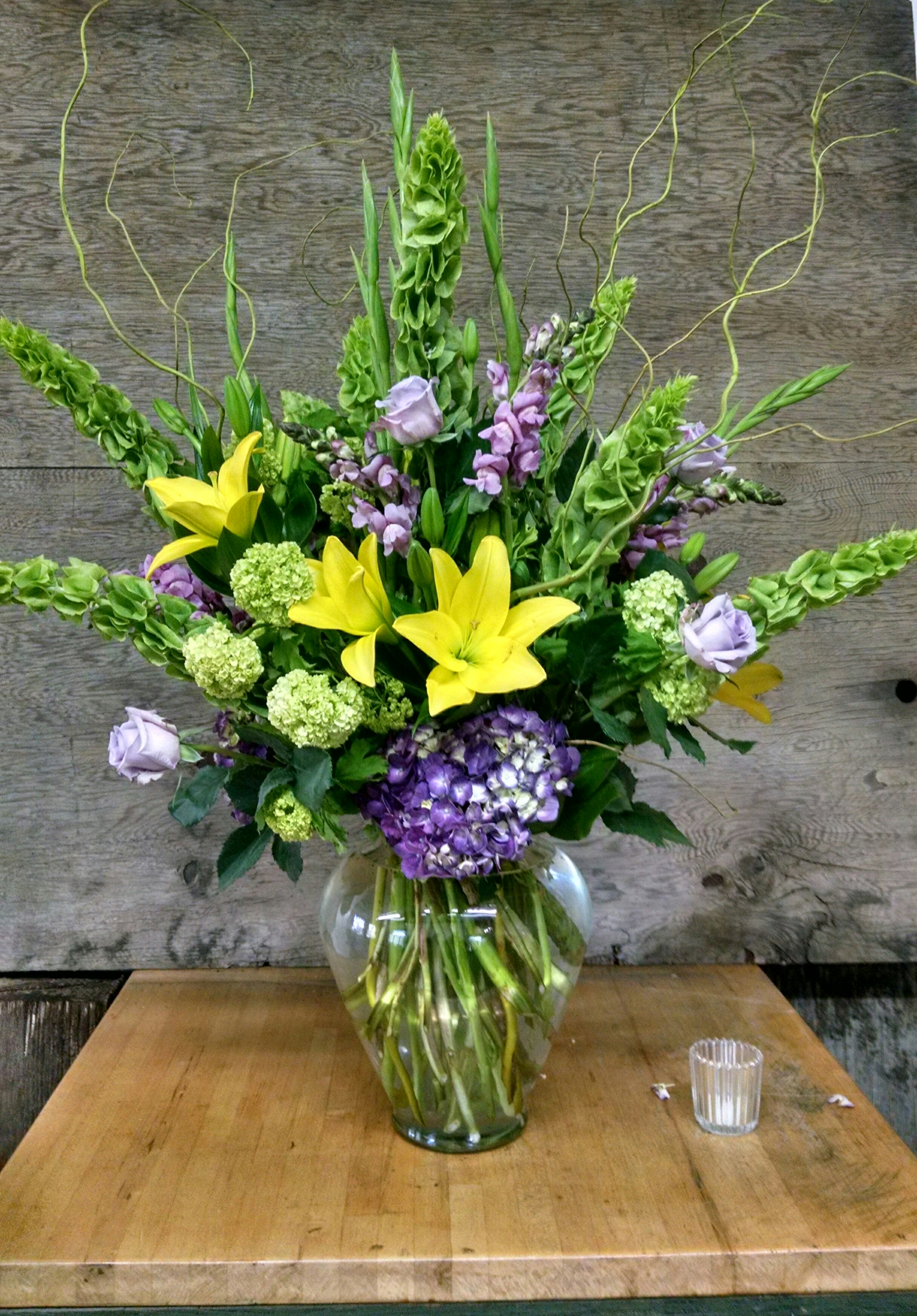 Tall bouquet of yellow lilies, purple blooms, and green flowers in a glass vase