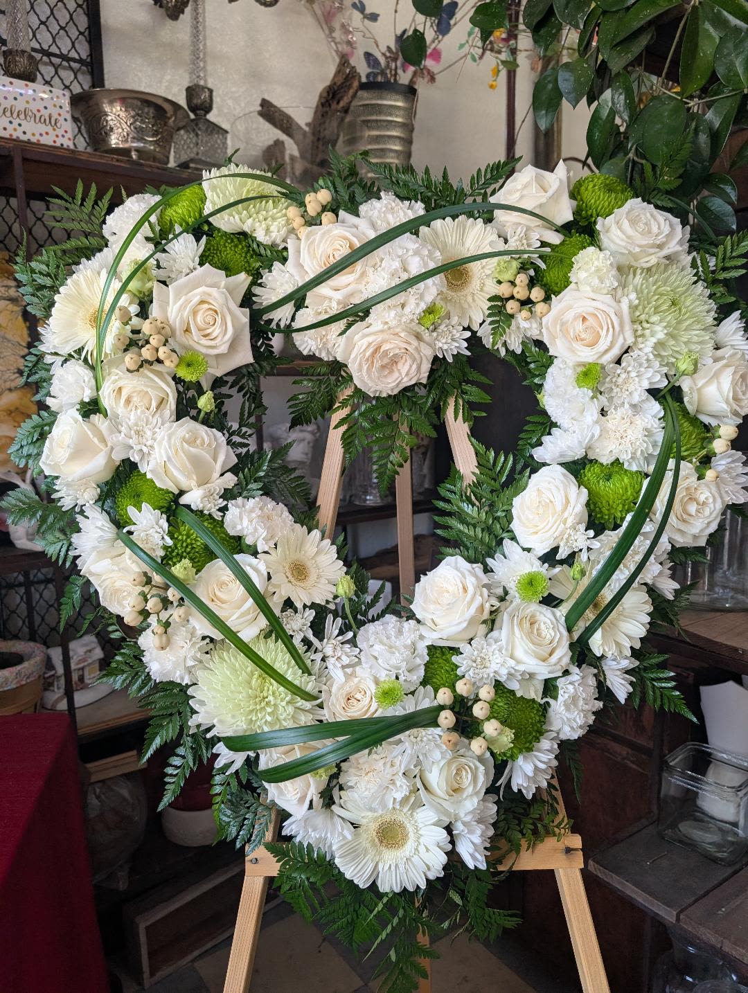 White and Green Heart - A lush heart-shaped sympathy arrangement that speaks to quiet remembrance, arranged on a wooden easel for memorial services or graveside tributes. The white and green heart uses classic white roses, white gerbera-style daisies, and round white and green chrysanthemums to create a soft, unified face; the form and color make it an appropriate choice for funeral or sympathy occasions while still feeling tender and sincere.  Tightly clustered blooms form the large heart, with white carnations and mixed white-and-green varieties filling the shape for a full, polished look. Long curved blades of greenery sweep across the front to add graceful movement and frame the heart, while decorative berries and dark foliage provide subtle contrast and depth. The arrangement reads formal and refined, with the wooden easel giving it a dignified, upright presentation suitable for viewing in a service setting.  Choose this piece to offer comfort to a family or to honor the memory of someone who loved classic, understated flowers-the heart shape delivers a message of love while the white palette conveys respect and peace. Sized to make a strong visual statement at a service, the easel-mounted display arrives ready to place; minimal care is needed beyond keeping blooms cool and sheltered until the memorial.
