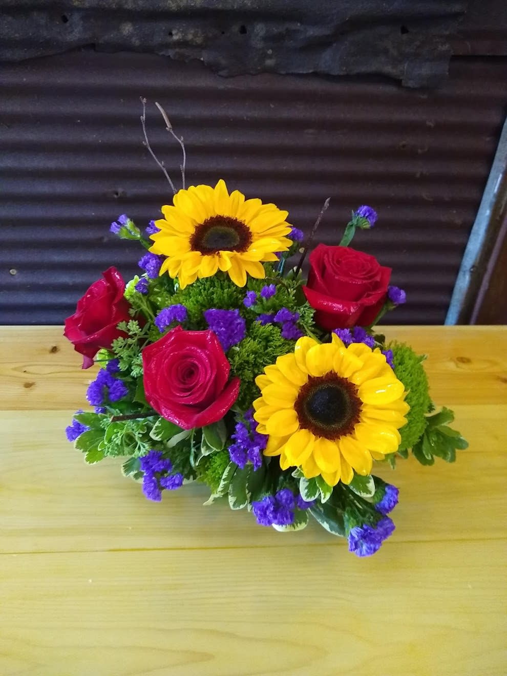 Falling For You - A sweet, petite dish of fresh sunflowers, red roses, mixed greens and purple statice. A perfect pick me up. Just the right size to say Hi Or I have been thinking about you.   One sided fresh arrangement designed in a plastic dish container. 