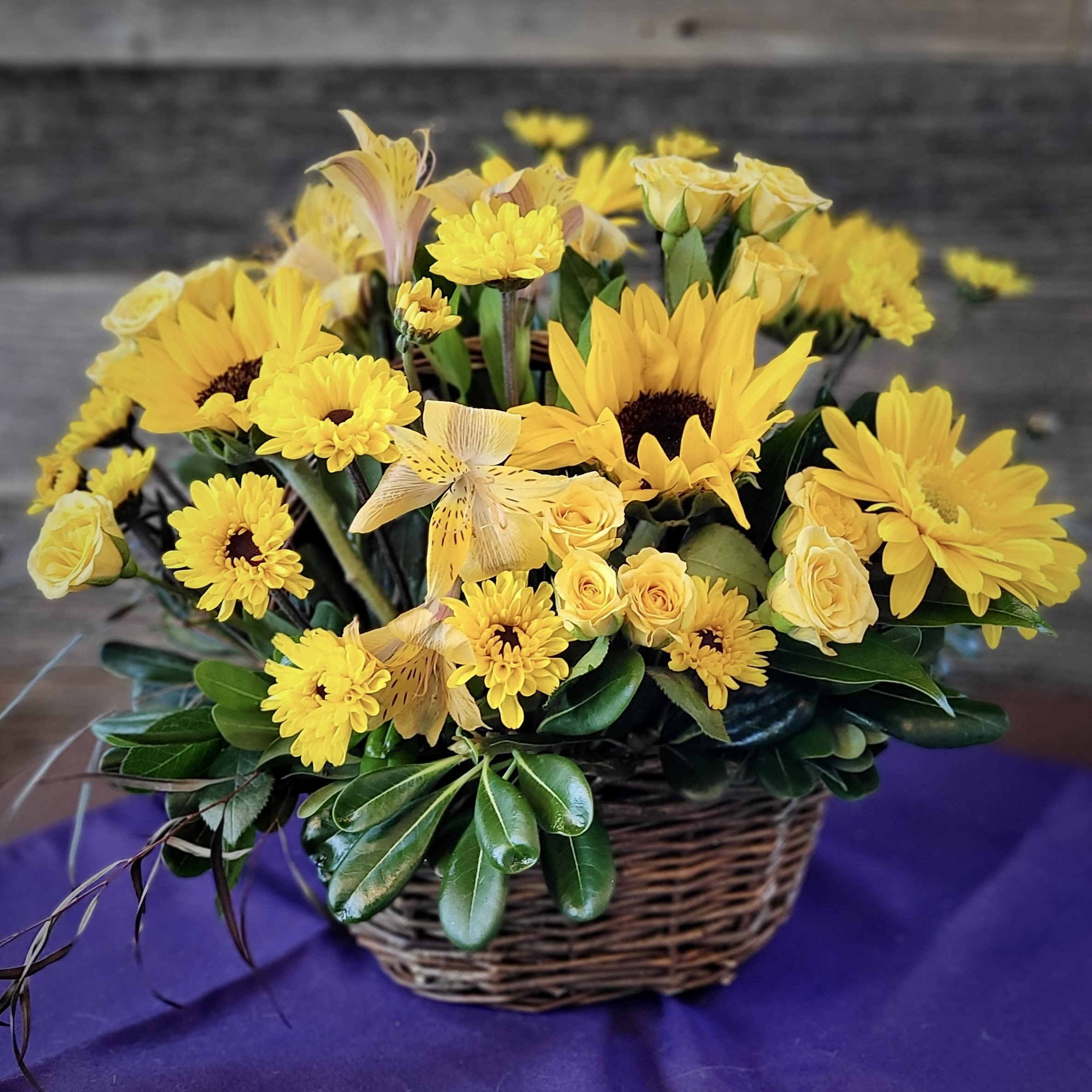 Basket arrangement of yellow sunflowers, roses, mums, and daisies with glossy foliage.