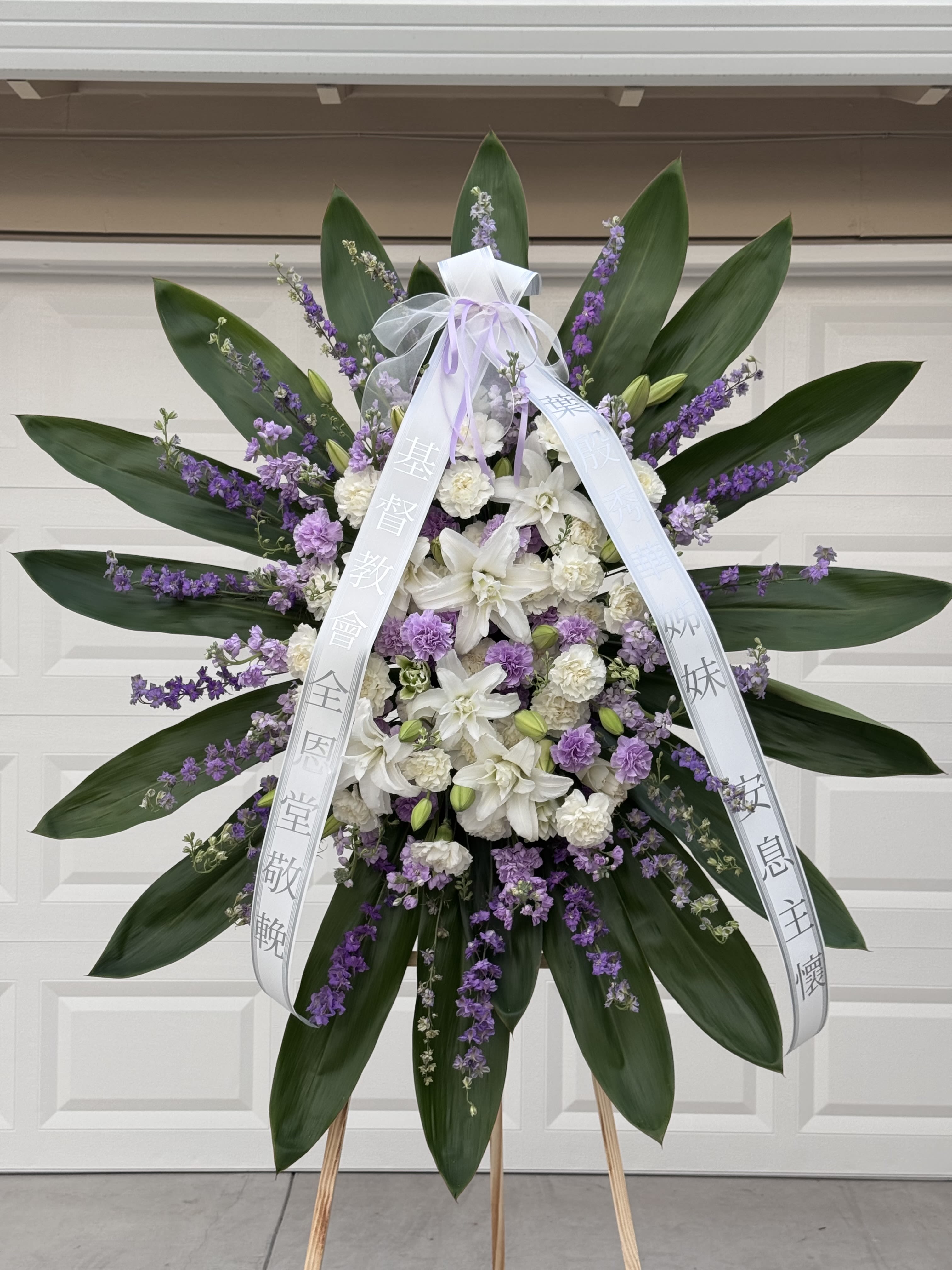 White Lilies and Purple Carnations Standing Spray - Include: - 1 wooden stand 66 inches - 2 free Banners (Max 8 Words each Banner) To Add BANNER MESSAGES ===Please Copy and Paste Below into "Special Instruction" box === Banner 1: (message here) Banner 2: (message here)  *****Please note that this wreath has to order in advance 2 days before delivery date.*****