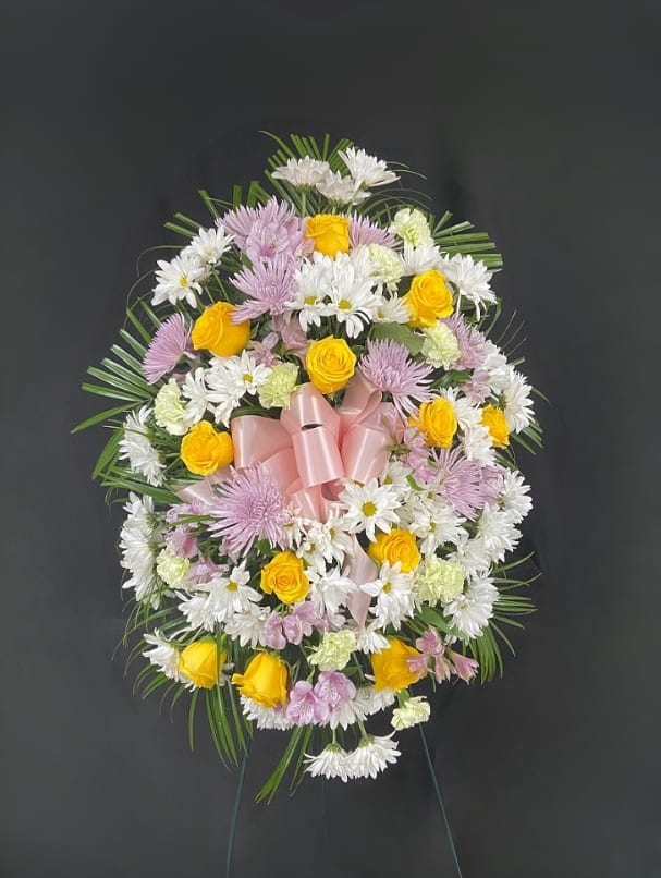 Standing floral wreath with yellow roses, white daisies, and pink ribbon