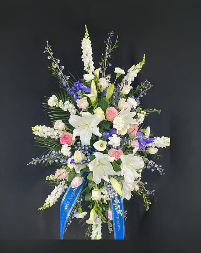 Skies Above Standing Spray - When sorrow comes, look to the skies above for comfort. This beautiful standing spray includes white Oriental lilies, blue delphinium, blue iris (seasonal), white mini calla lilies (when available), pink roses, white snap dragons, white stock, and white carnations. Arrangement is mounted on a 4.5ft standing easel for prominent display at funeral services.