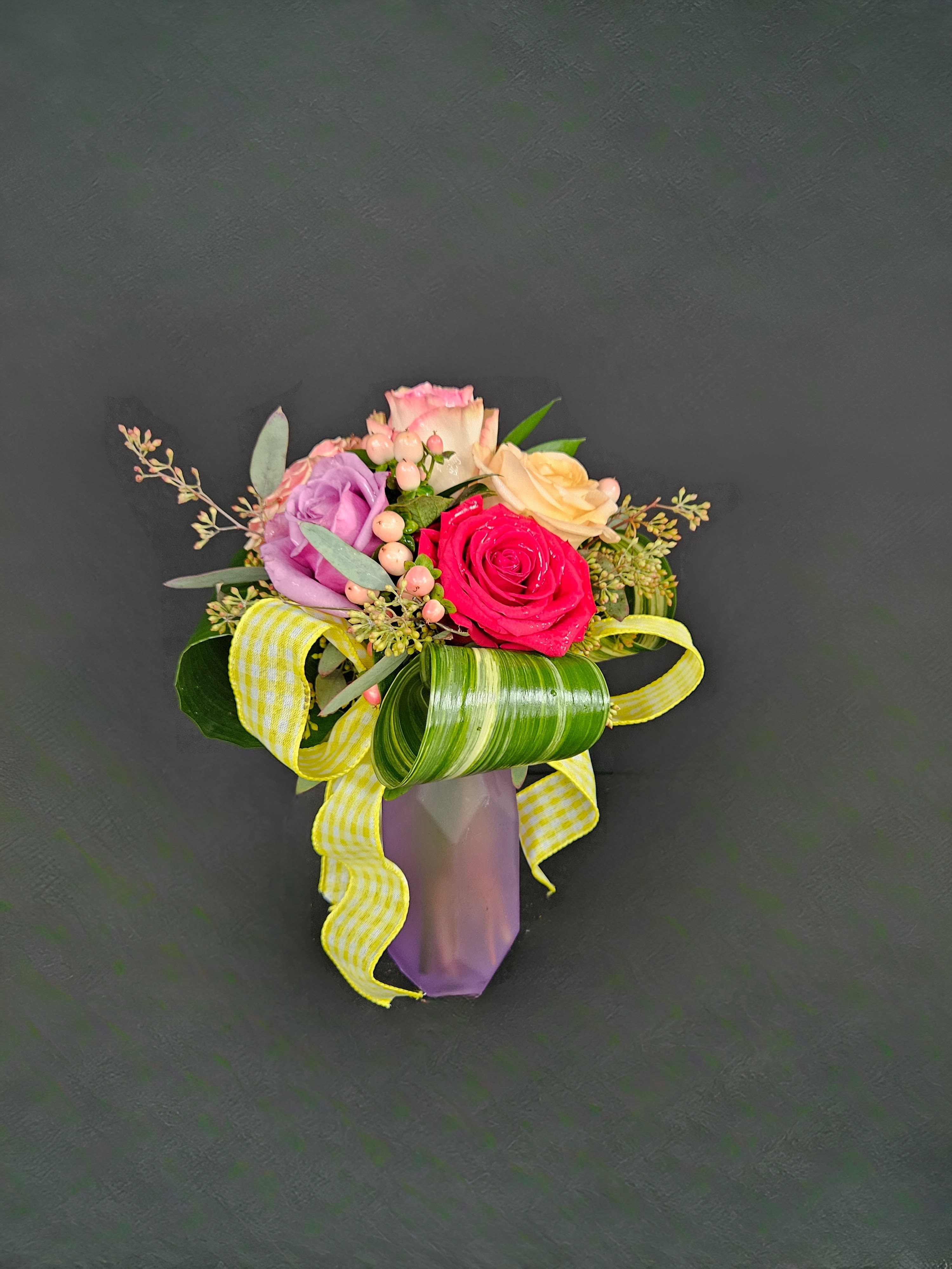 Small arrangement of multicolored roses with berries and yellow ribbon in a purple vase