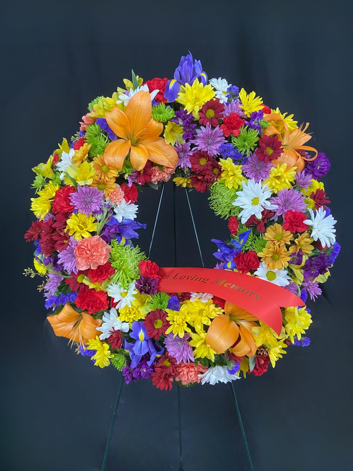 Lovely Memories Standing Wreath - Vibrant and full just like the life they lived. This wreath is mounted in a 54" easel and packed with color. Roses, carnations, lilies, iris, mums, alstro, and statice shine through with bright reds, blues and yellows against a backdrop of white and green. Includes 1 custom printed banner.