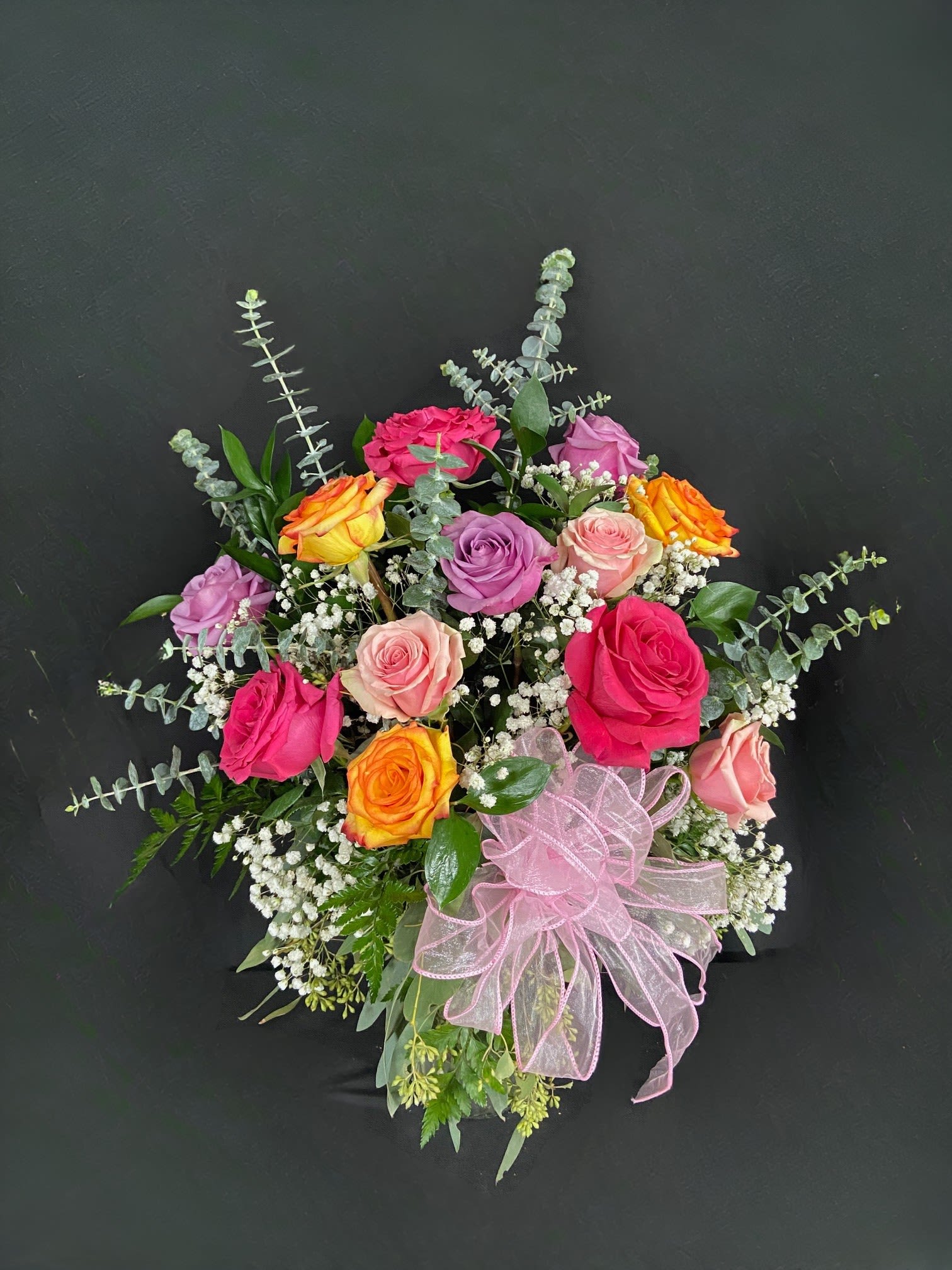 Mixed arrangement of pink, peach, lavender, and yellow roses with white filler flowers and a pink ribbon bow