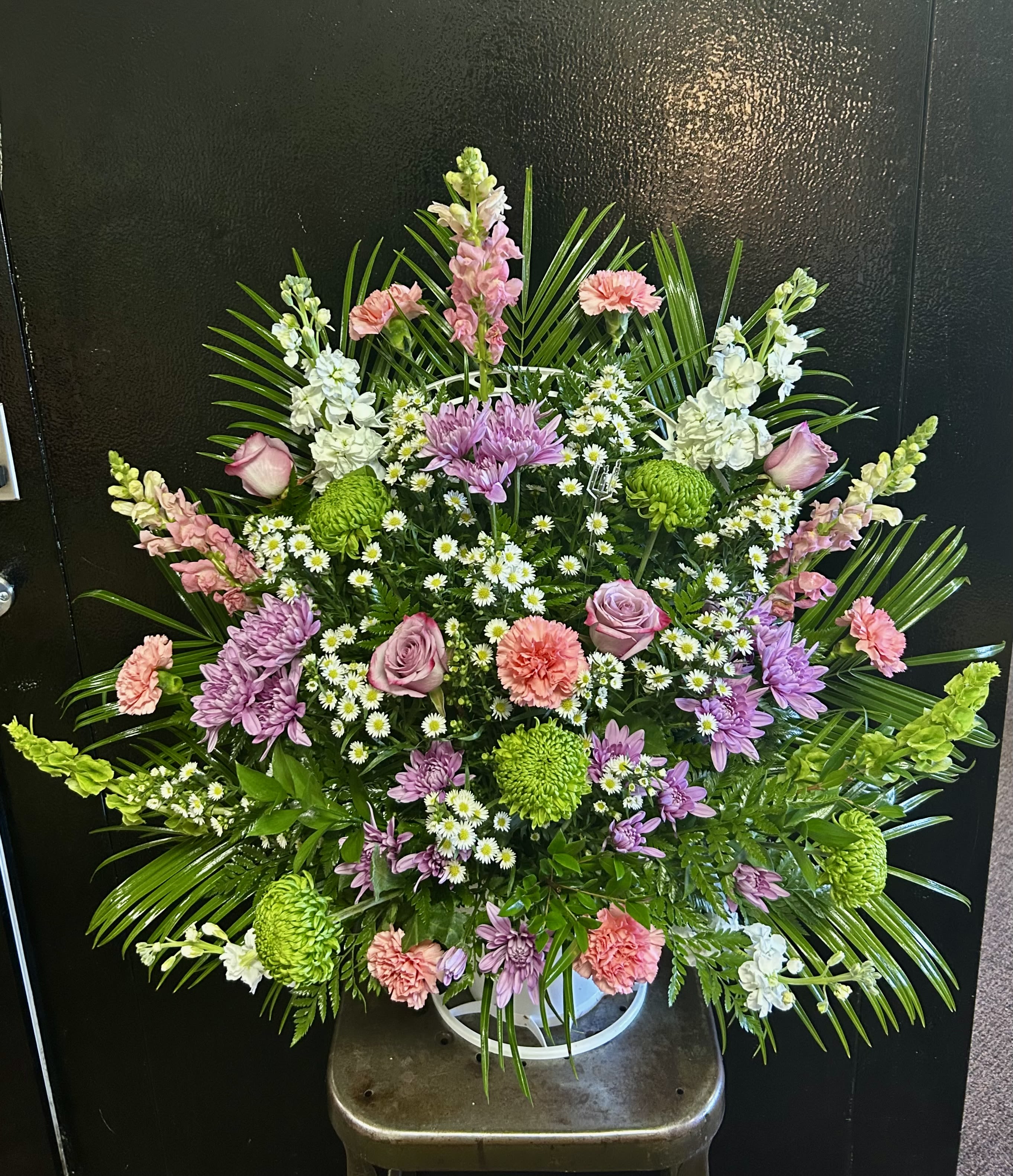 Beloved Memories Traditional Sympathy Basket - A soft, uplifting sympathy basket designed to comfort and honor - this large upright flower arrangement brings together pink roses, purple and white chrysanthemums, cheerful daisy-like blooms, and ruffled carnations to create a serene, memorable tribute. The fan-shaped silhouette, accented by tall snapdragon spikes and broad palm fronds, reads as both formal and gently hopeful, making it well suited for sympathy services or as a thoughtful remembrance piece for the person you're thinking of.  Set in a white plastic container for stability, the design is dense and full with bright green accents that add freshness to the pink, purple, and white palette. The layered textures - silky rose petals, feathery mums, crisp snapdragon spikes, and soft carnation ruffles - create visual depth whether placed beside a casket or displayed at a memorial service. Sized to make a strong, polished statement, this arrangement offers a graceful way to express caring, sympathy, and lasting memories.
