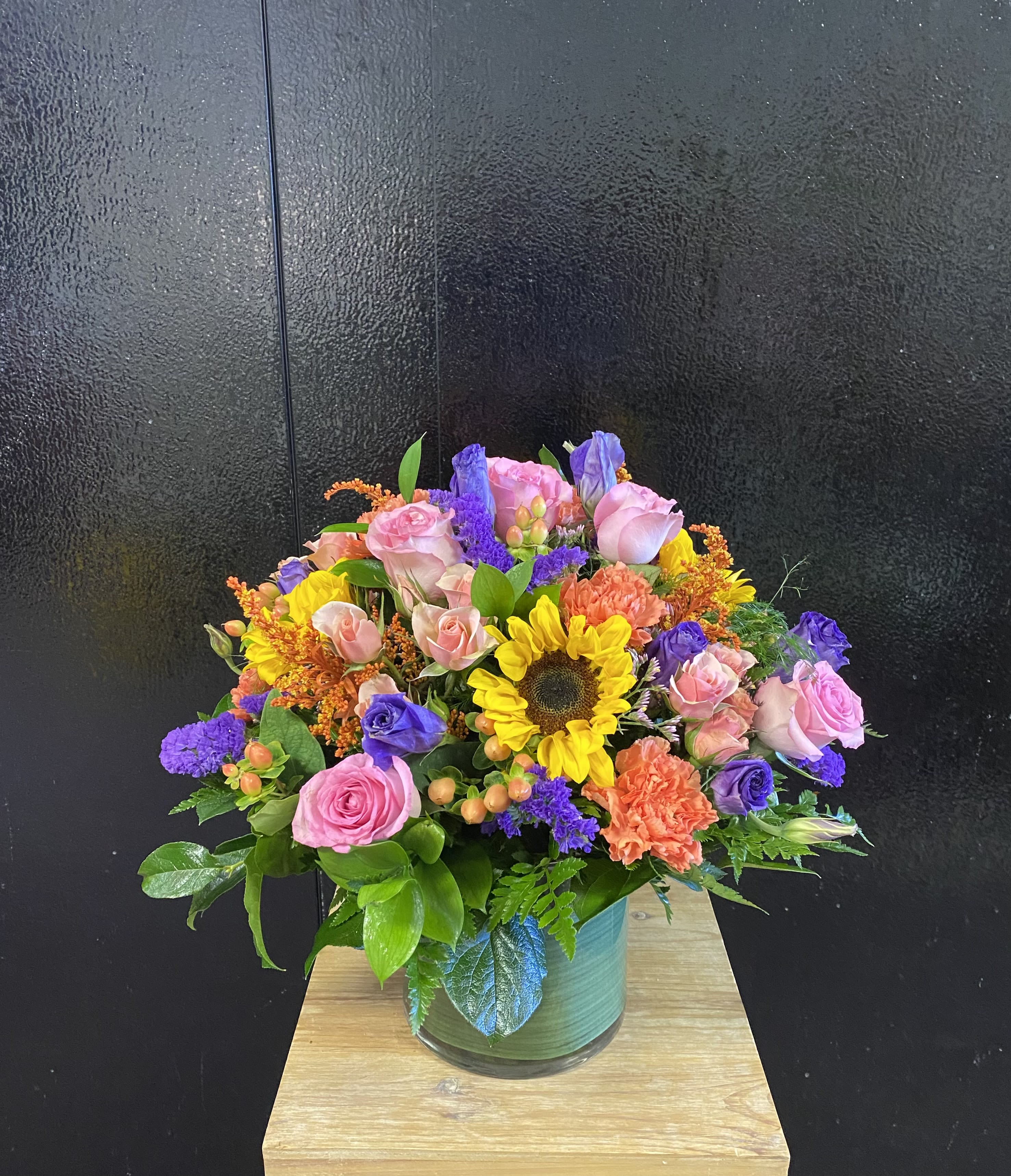 Joyful Blooms Bouquet - A lush, rounded bouquet of pink roses paired with bright yellow sunflowers creates an immediate burst of joy-perfect as a birthday bouquet or a cheerful congratulations arrangement. The mix of orange carnations and purple filler flowers lifts the palette, so the person you're thinking of will feel genuinely celebrated the moment the blooms arrive.  Packed closely for a full, layered look, this flower arrangement sits in a clear glass vase that gives the design a polished, everyday elegance. Expect a dense composition of roses, sunflowers, carnations and complementary seasonal blooms in pink, purple, yellow, and orange tones-designed to read as vibrant from every angle and brighten any tabletop or office.  Send this as a feel-good gift to mark birthdays, promotions, new homes, or "just because" moments; it reads as lively and upbeat while still being refined. The vase is included and the arrangement is sized to make an immediate visual impact without overwhelming smaller spaces-simple care: keep fresh water, trim stems occasionally, and display out of direct heat for the longest enjoyment.