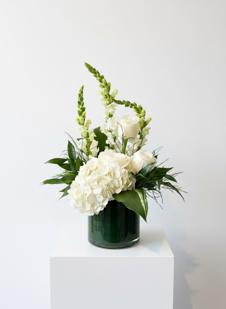 Sympathy Vase Arrangement - A classic sympathy arrangement in a vase is a timeless and elegant expression of remembrance and respect. Designed with a serene palette of white, green, and soft neutral tones, this arrangement often features graceful blooms such as roses, lilies, hydrangea, and seasonal accents, complemented by fresh greenery for a natural, calming look.  Arranged in a clear or understated vase, its balanced and refined style makes it appropriate for both funeral services and delivery to the home of loved ones. The gentle color palette symbolizes peace, purity, and comfort, offering a quiet tribute that honors a life beautifully lived.