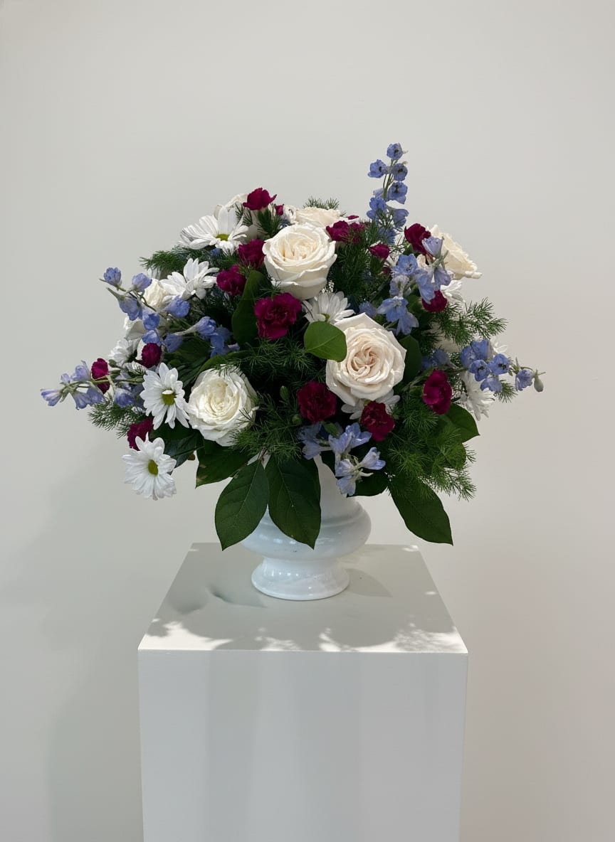 Arrangement of white roses, white daisies, burgundy carnations, and blue delphinium in a white pedestal vase