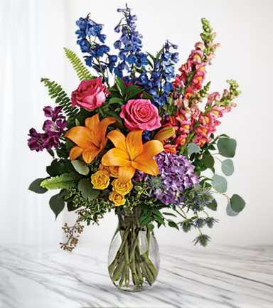 Colorful bouquet with lilies, roses, and tall blue and pink blooms in a glass vase