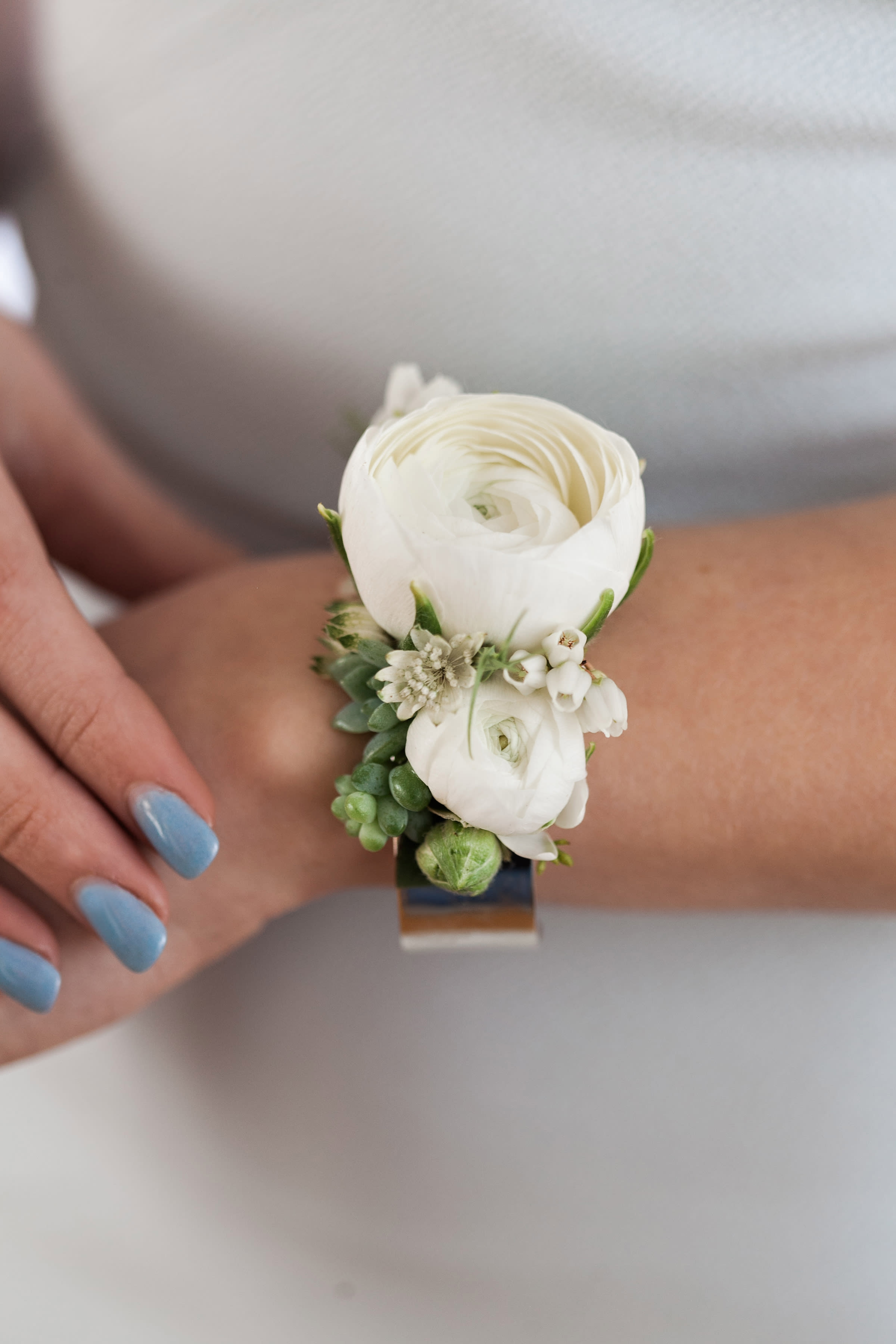 Ranunculus Corsage on a Silver Cuff - Pick Up Only - Cuff corsage featuring ranunculus with bits of foliage and other filler flowers. Please note in the 'Special Instructions' box what color you'd like the focal flower to be and any other accent colors you'd like to see in the design. Please note that each corsage is botanical, unique and may differ from the image shown online. If no colors are noted in the 'Special Instructions' then we will use white flowers.  Please Note: The high shine silver cuff is currently unavailable, we are using a silver cuff that is matte.   Please note that corsages and boutonnieres are non-refundable after being made. 