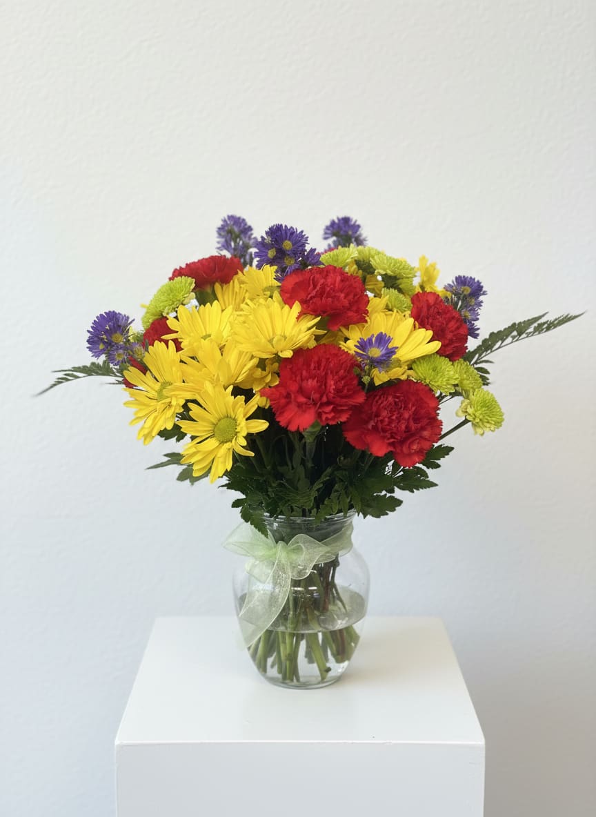 Sun and Cheer Bouquet  - Sun-soaked yellow daisies take center stage in this flower arrangement, a joyful bouquet perfect for a birthday, spring celebration, or a bright "just because" surprise. The mix of red carnations and small purple asters creates lively contrast that will make the person you're thinking of feel genuinely celebrated and noticed.  Crafted into a rounded, casual bouquet, the blooms sit in a clear glass vase tied with a pale green ribbon for an uplifting, gift-ready presentation. Expect cheerful yellow daisies, clustered red carnations, and delicate purple asters with fresh green accents-an arrangement that reads warm, energetic, and effortless.  Ideal for sending thanks, sending smiles, or marking a happy milestone, this size is easy to display on a kitchen table or desk and requires simple care: fresh water in the vase and a cool spot out of direct afternoon sun will keep the colors bright. Order with confidence for a cheerful surprise that reads as thoughtful and spontaneous.