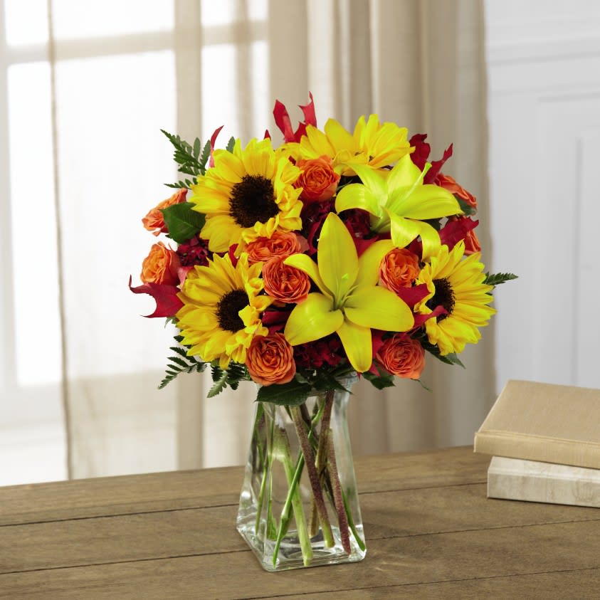 The FTD Harvest Heartstrings Bouquet - The FTD® Harvest Heartstrings™ Bouquet brings sunlit autumn beauty straight to their door. Unforgettable mini sunflowers catch the eye at every turn surrounded by yellow Asiatic lilies, red dianthus, orange spray roses and lush greens to create a stunning fresh flower arrangement. Presented in a clear glass gathered square vase and accented throughout with red glycerized oak leaves, this flower bouquet is set to make an excellent birthday, thank you, get well or Thanksgiving gift. 