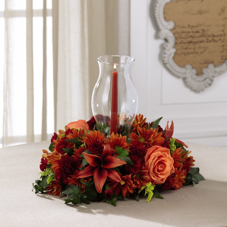 The FTD Heart of the Harvest Centerpiece - The FTD Heart of the Harvest Centerpiece exudes warmth and autumn elegance to create the perfect addition to their fall table. Orange roses, burgundy carnations, rust Asiatic lilies, burgundy mini carnations, bronze traditional daisies, yellow solidago and lush greens are beautifully arranged to surround a clear glass hurricane holding a single burgundy taper candle to create an eye-catching centerpiece. Set to bring color, life and light to your special recipient's gathering, this fresh floral centerpiece will create an excellent Thanksgiving gift. 