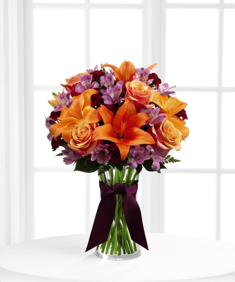 FTD Harvest Heartstrings Bouquet - The FTD Harvest Heartstrings Bouquet is a fresh burst of color and autumn light that will surround your special recipient with beauty. Exquisite Cherry Brandy Roses are brought together with purple Peruvian Lilies, orange Asiatic Lilies and burgundy mini carnations accented with lush greens and arranged in a classic clear glass vase. Highlighted by a deep eggplant satin ribbon tied around the neck of the vase, this bouquet is an arrangement that speaks to the heart of the fall season. 