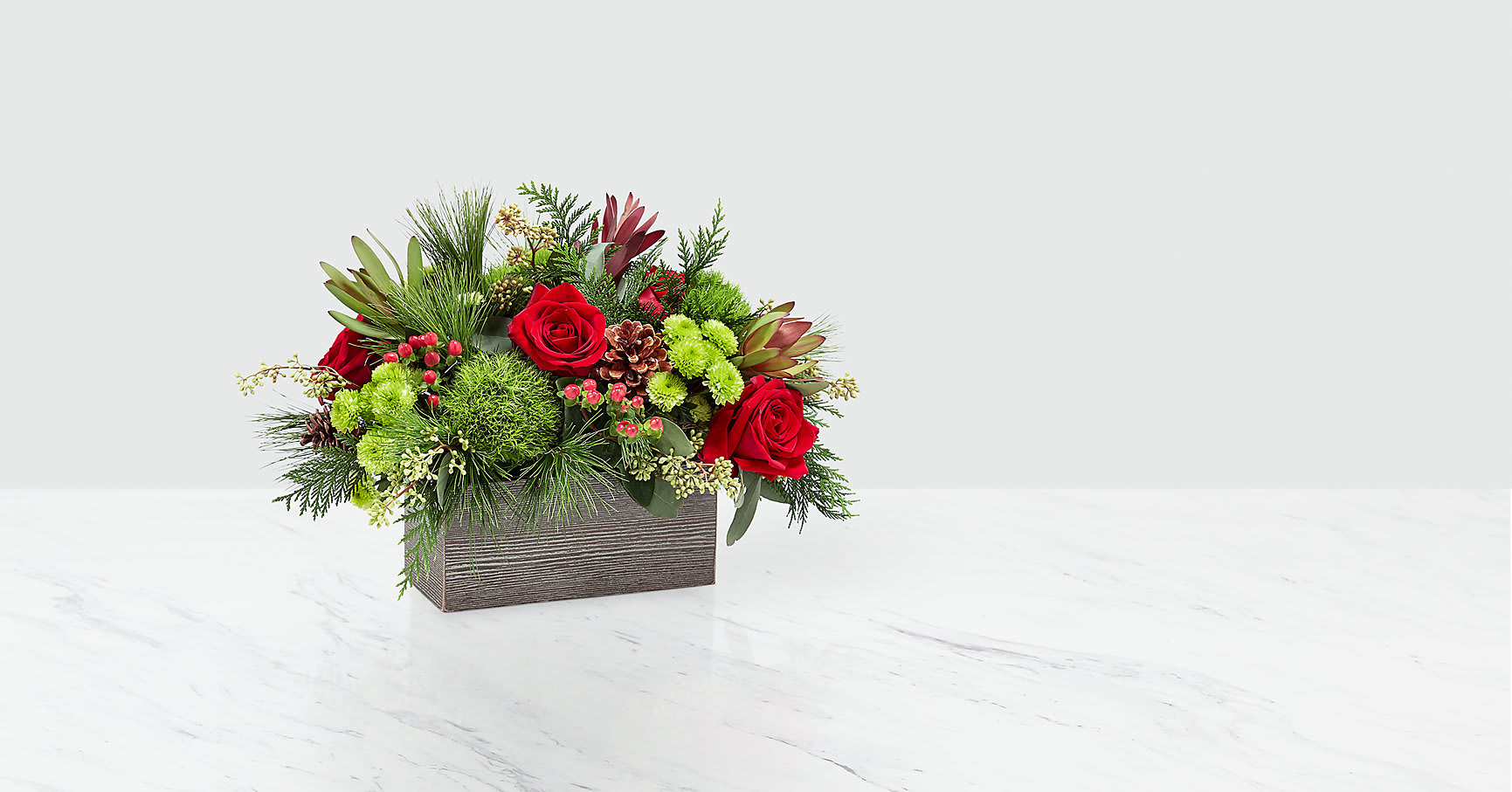 Christmas Cabin™ Bouquet - This stunning bouquet is handcrafted to bring the cozy feel of the holidays into your home. An array of red roses, green button pompons, and seeded eucalyptus is set in a weathered, wooden rectangular box and can make any morning feel like a cozy Christmas morning. The protea, berries and pinecones add a finishing touch.   ITEM #18-C7D
