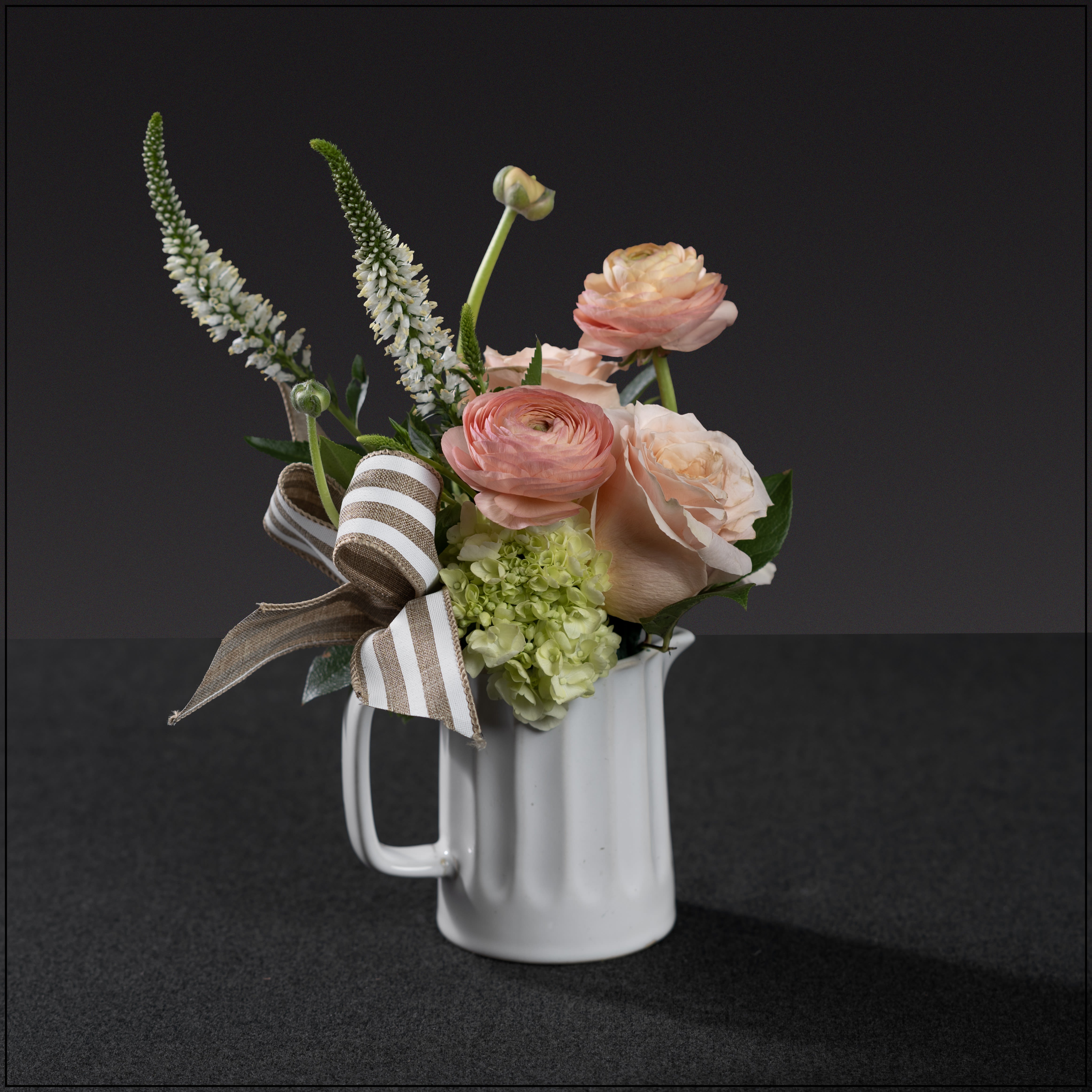 Mother's Day Whimsy - A petite white pitcher with a subtle mix of roses, miniature hydrangea, ranunculus, veronica, and a vintage carnation, accented with a beige and white striped bow. A lovely gift for any occasion. Approximately 13" tall and 7" wide.