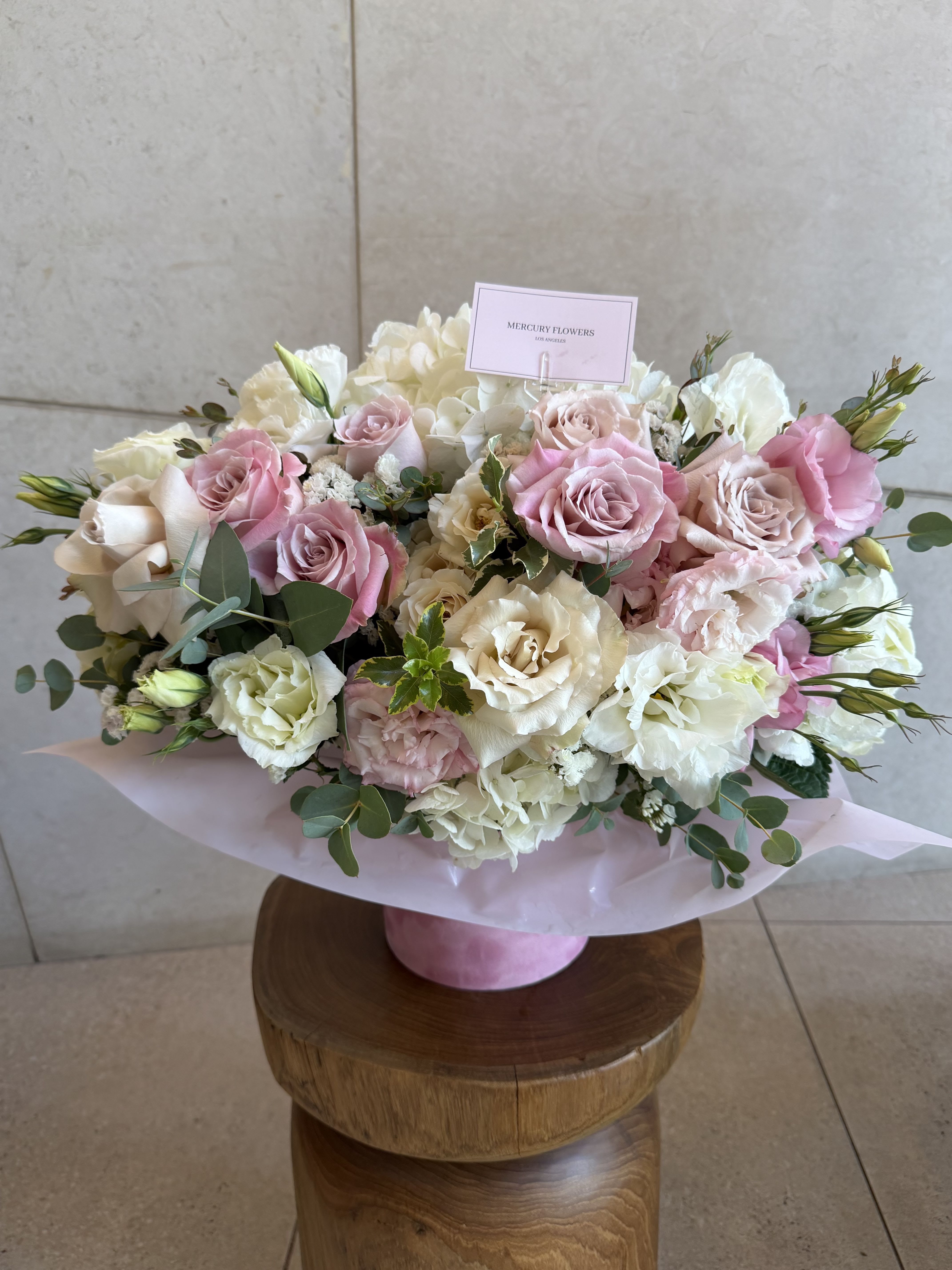 Miss Dior box - A soft, rounded bouquet in a pink hatbox that reads like a romantic note: blush pink and ivory roses mingle with ruffled white and pale pink lisianthus, creamy hydrangea clusters, and airy eucalyptus for a refined, pastel arrangement perfect for an anniversary bouquet, Mother's Day gift, birthday surprise, or a "thinking of you" moment. The presentation - a pale pink cylindrical box with matching floral wrap and a tucked branded card - makes it feel gift-ready the moment it's delivered.    This full, low arrangement is densely clustered for an elegant, tactile look; large garden roses provide focal blooms while lisianthus and hydrangeas add ruffled texture and volume. The palette of pink, white, green and soft peach creates a gentle, celebratory mood that suits weddings, Valentine's Day, graduations, housewarmings, and sympathy or get-well wishes without feeling seasonal. Includes a clear card holder and display card for your message, and arrives wrapped in pale pink paper inside the pink hatbox for an instant, polished presentation-ideal for anyone you want to make feel truly seen.