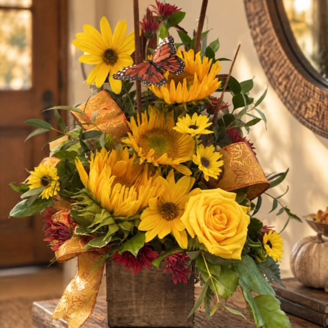Yellow sunflowers and roses arranged in a rustic vase with ribbon and a butterfly pick.