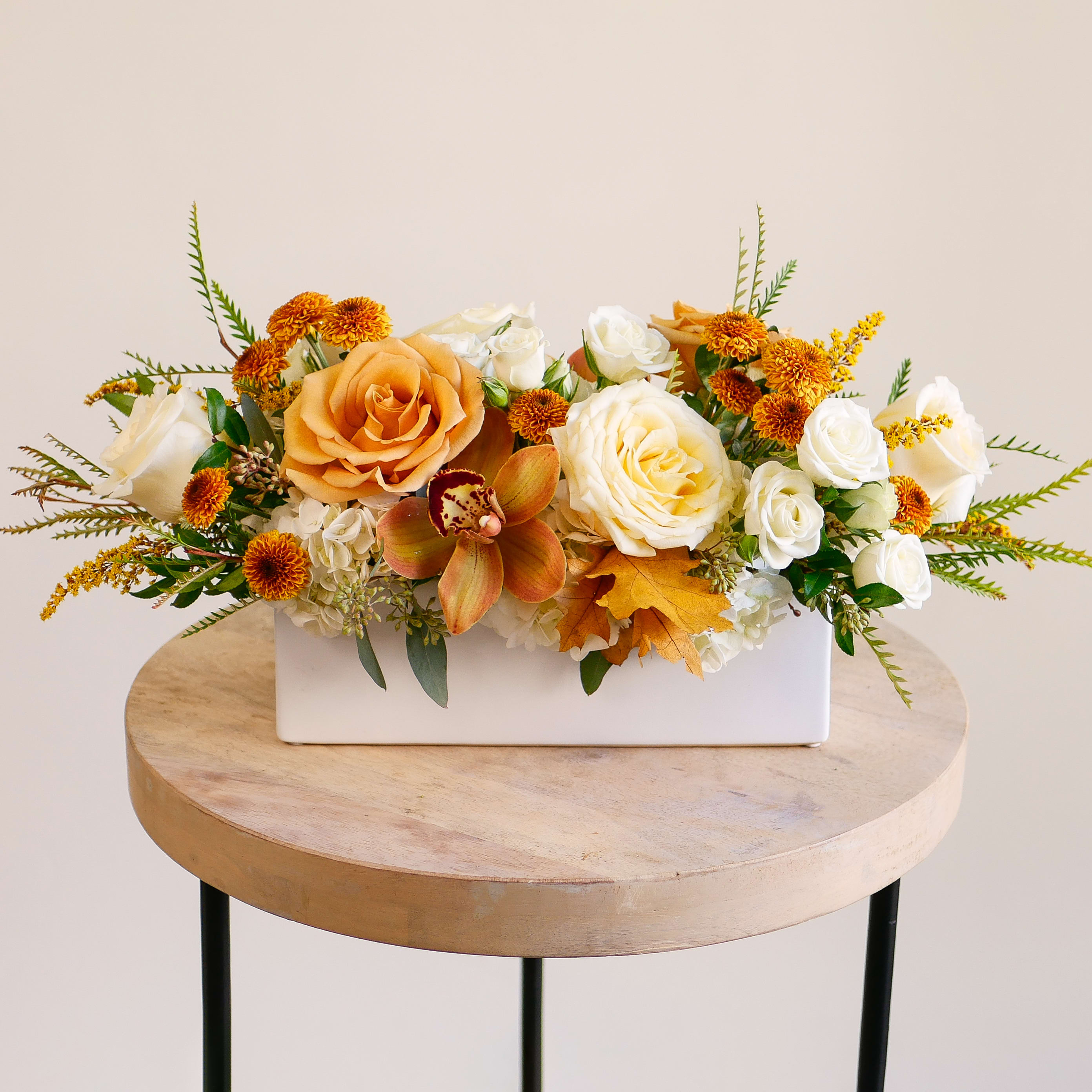 Savanna Centerpiece - Arrangement approximately measures 8 inches tall and 18 inches long with flowers.  Celebrate the season with our breathtaking Savanna Centerpiece Flower Arrangement, a luxurious centerpiece that perfectly captures the warm, muted tones of autumn. This unique floral design features a harmonious blend of textures and colors, artfully arranged in a contemporary white rectangular vase.  Inside, you'll find elegant white and cream roses, deep golden and butterscotch garden roses, and delicate clusters of peachy, blush dahlias. The focal point is a striking burnt orange or rust-colored cymbidium orchid, lending an exotic, sophisticated flair. Accents of white hydrangeas, wispy solidaster, and dark, aromatic eucalyptus greenery complete the look, creating a rich tapestry of autumn flowers.  Whether you're looking for a stunning Thanksgiving centerpiece, a unique host or hostess gift, or simply want to bring the beauty of the Golden Hour into your home, this arrangement is the ideal choice. It’s perfect for a dining table, mantle, or entryway. Order your premium fall arrangement today!