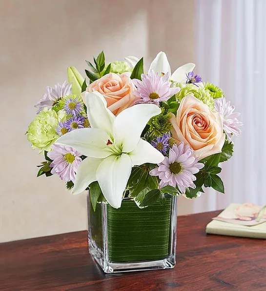 Mixed bouquet with white lilies, peach roses, and lavender daisies in a square glass vase