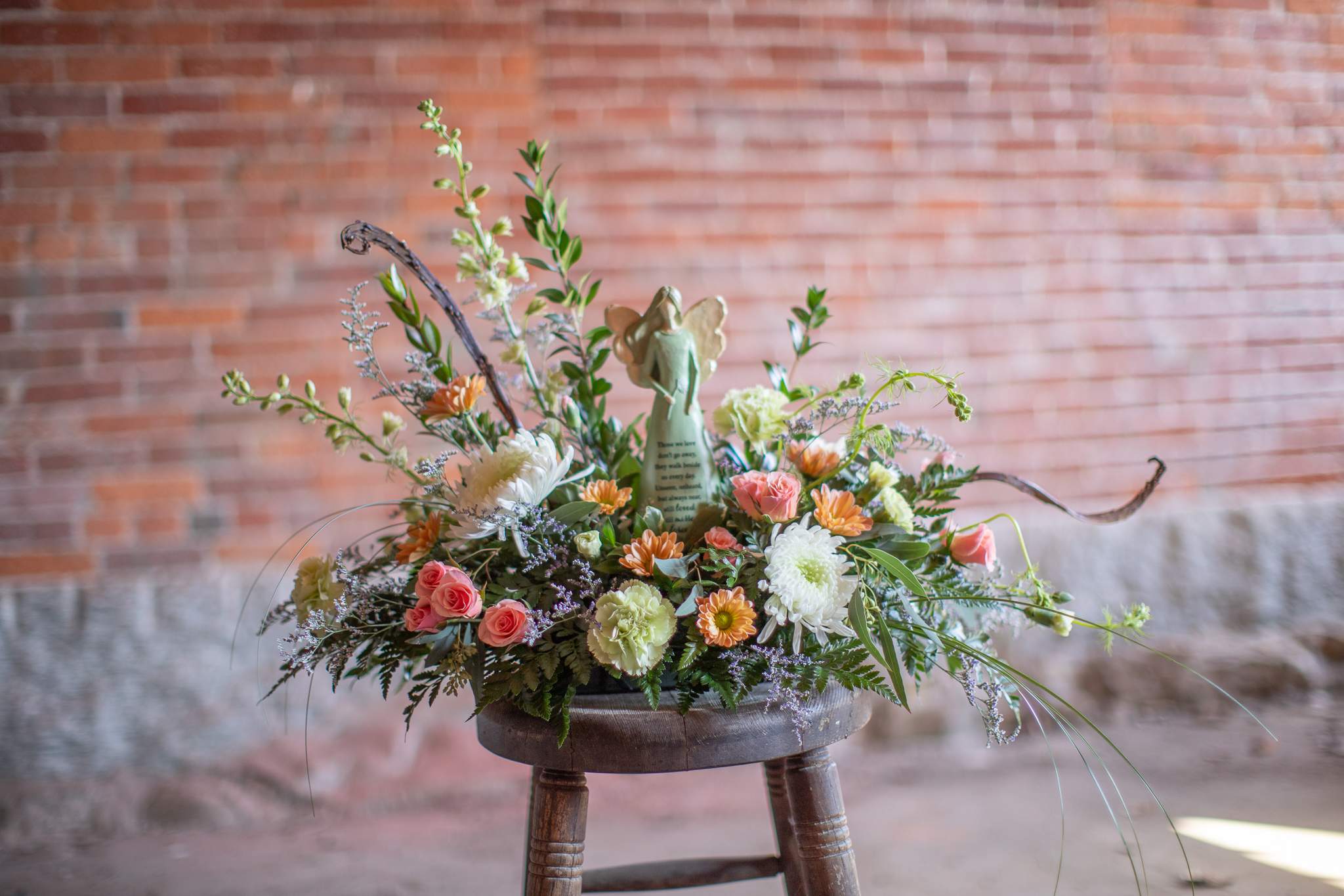 Low pastel arrangement of roses, mums, and daisies with an angel figurine in the center on a rustic stool