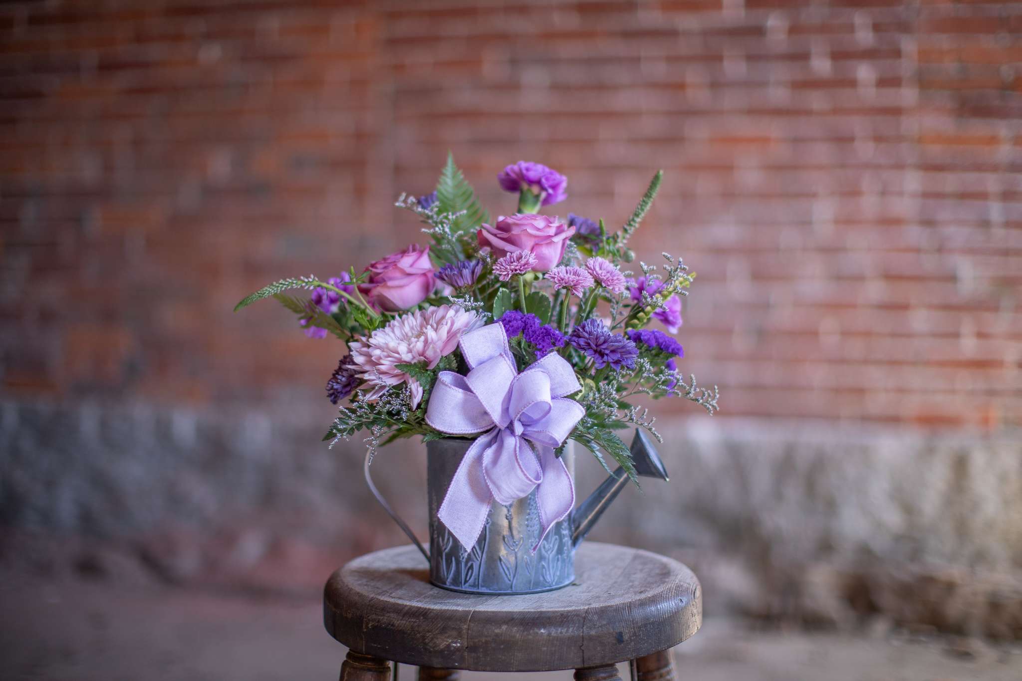 Purple and pink flower arrangement in a small metal watering can with a lavender bow