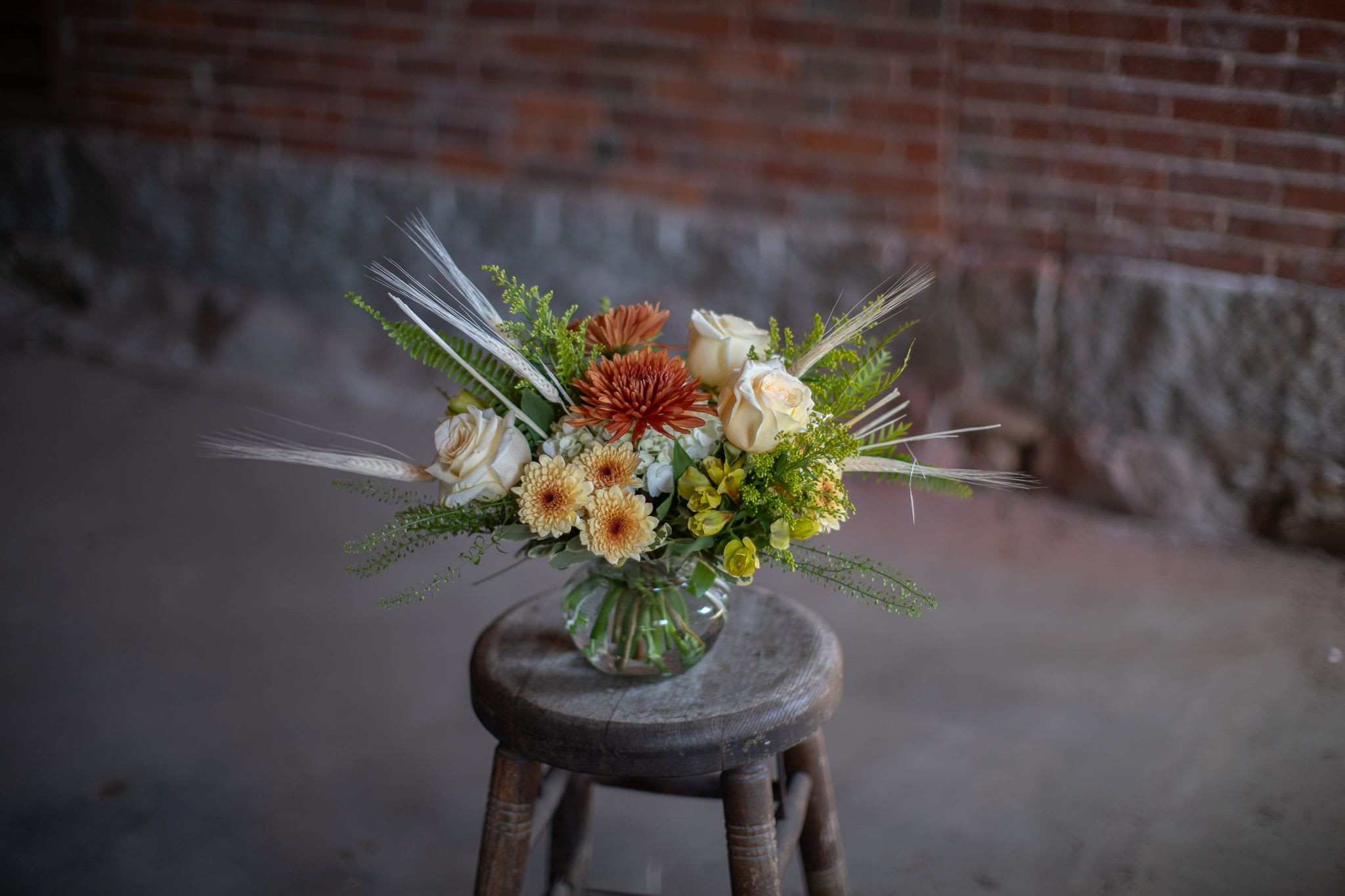 Fall Whisper - Wonderful bleached wheat whisps from pretty vintage-toned fall blooms in a shallow fluted recycled glass bowl style vase. Soft white hydrangea cradles creme brulé cushion pom pons or butterscotch toned spray mums. Soft cream or white roses. The design is two sided and more oblong style. Perfect for a centerpiece on a table or coffee table. Copper or bronze mums complete the look. Possible substitutions: In some instances we may use orange, cream, white or yellow alstroemeria, we may use butterscotch, peach or white spray chrysanthemums. We may also use mini green hydrangea (3) in place of the 1 white hydrangea photographed. White roses, peach, cream, or yellow roses may be used. Overall, the look and feel will be the same. Please call with questions about substitutions.   