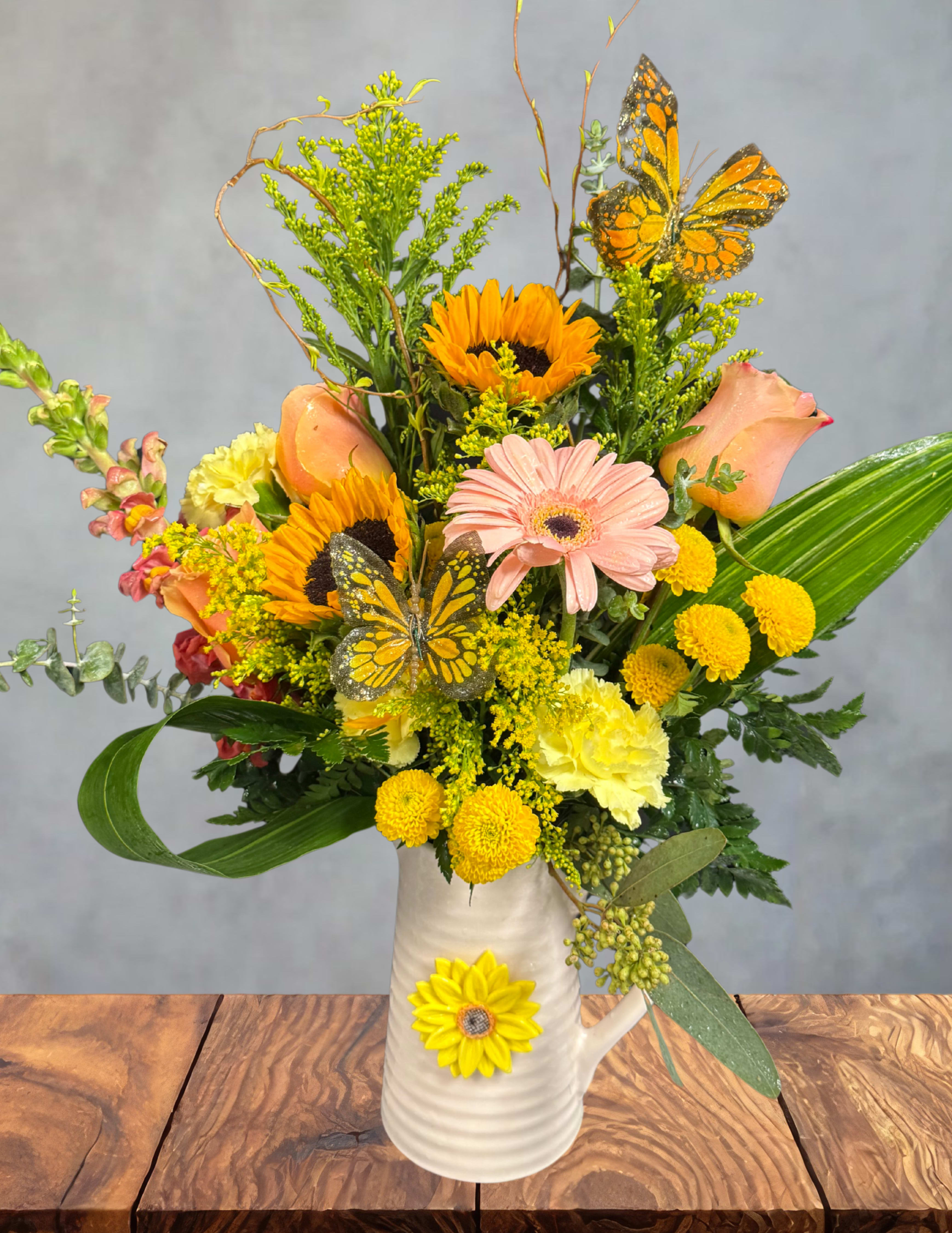 Butterfly Morning -  Butterfly Morning A warm, rustic embrace of sunshine and gentle beauty. Butterfly Morning features radiant sunflowers, glowing roses, cheerful daisies, and golden accents arranged in a charming ceramic pitcher adorned with a sunflower motif. Two graceful monarch butterflies perch among the blooms, creating the feeling of a peaceful morning garden coming to life.  Comforting, bright, and full of heartfelt warmth, this design is ideal for birthdays, thank‑yous, or anyone who could use a little morning light in their day.