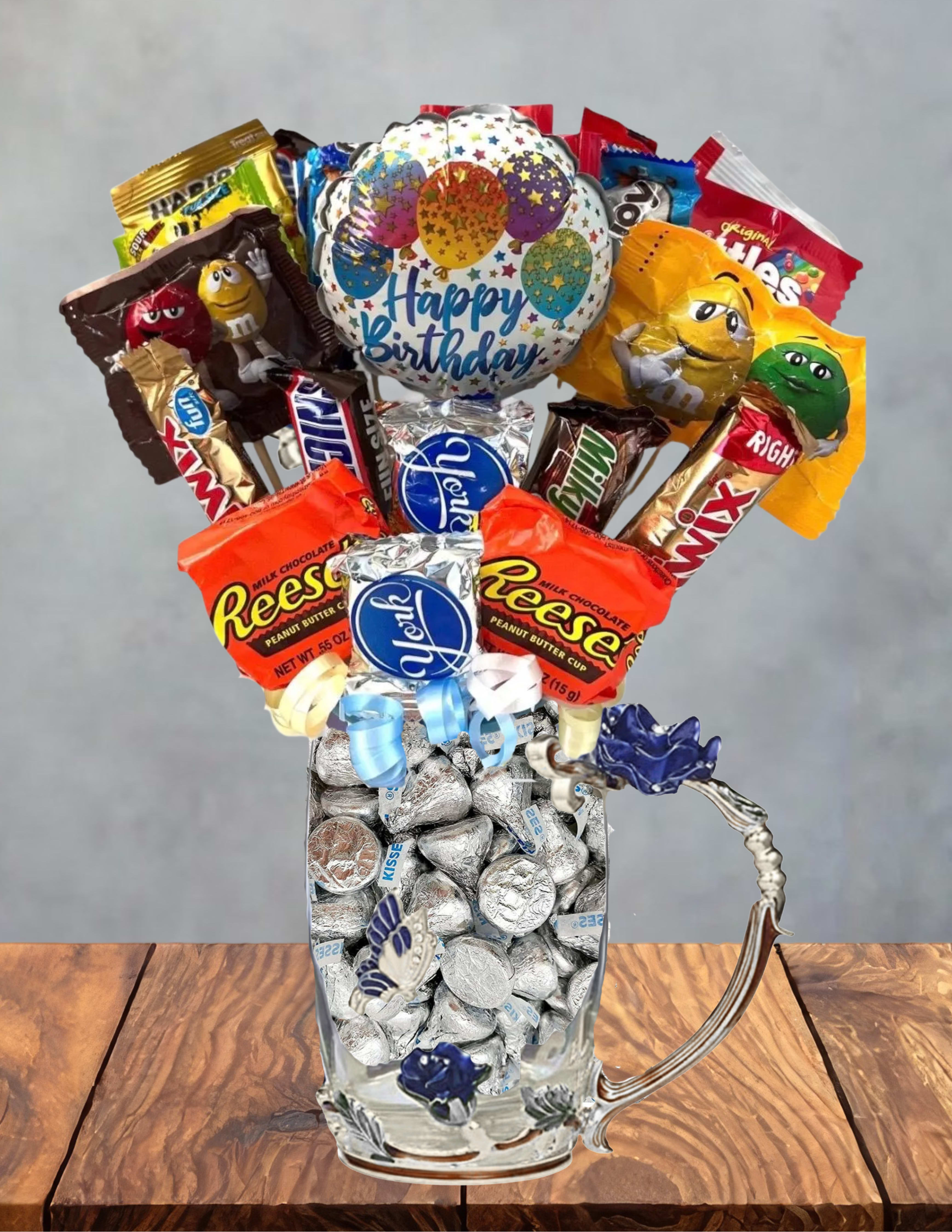 Blue Birthday candy bouquet in pretty tea glass - Bright, playful, and ready to celebrate-this Blue Birthday candy bouquet in a pretty tea glass makes a joyful birthday gift that will make the person you're thinking of grin. Designed as a birthday gift set rather than a floral arrangement, it features a round birthday balloon topped above the treats to create a festive focal point perfect for birthdays, milestone celebrations, or a cheerful pick-me-up.  Assorted candy bars and wrapped chocolates, including chocolate kisses, are artfully arranged in a clear glass mug so the colorful red, orange, blue, gold, and silver wrappers show through for instant visual impact. A decorative ribbon and a delicate butterfly decoration finish the presentation, turning simple sweets into a charming keepsake. The clear tea-style glass mug also serves as a reusable holder for desk supplies or future treats.  This is ideal for a coworker's desk surprise, a child's birthday, or anyone who loves sweets and bright, playful packaging-the kind of gift that makes someone feel seen and celebrated. Because it's a hard-good add-on, order now to include a balloon topper and ribbon; the arrangement ships ready to present with everything in place and requires no special care beyond keeping the chocolates in a cool, dry spot.