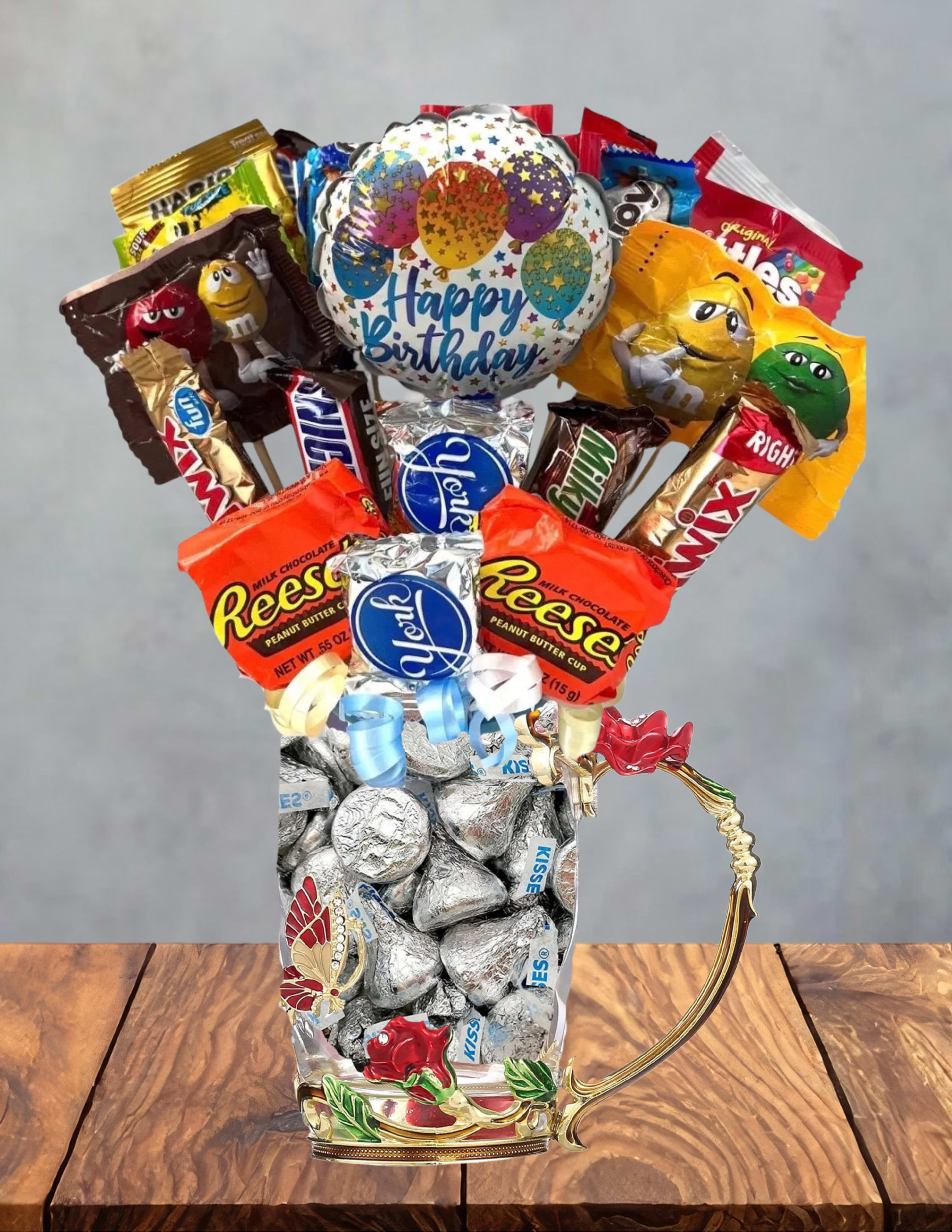Pink Birthday candy bouquet in pretty tea glass - A playful candy bouquet built from favorite sweets-this Pink Birthday candy bouquet in a pretty tea glass is a cheerful birthday gift designed to delight. The arrangement reads as a novelty birthday bouquet rather than floral, topped with a round "Happy Birthday" balloon and overflowing from a tall, tea-glass style mug to create an immediate celebratory focal point perfect for birthday parties, coworkers, or anyone who loves sweets.  Assorted candy bars and wrapped chocolates in mixed color packaging (red, orange, gold, blue, and silver) are densely arranged to look abundant and festive; ribbon accents finish the composition and keep everything looking polished. The decorative mug holds the pieces securely and doubles as a reusable keepsake, while the balloon adds height and instant party-ready flair. This ready-to-give candy bouquet is ideal for surprising the person you're celebrating-no watering or care required-just order with the date and we'll prepare it to arrive looking full and festive.