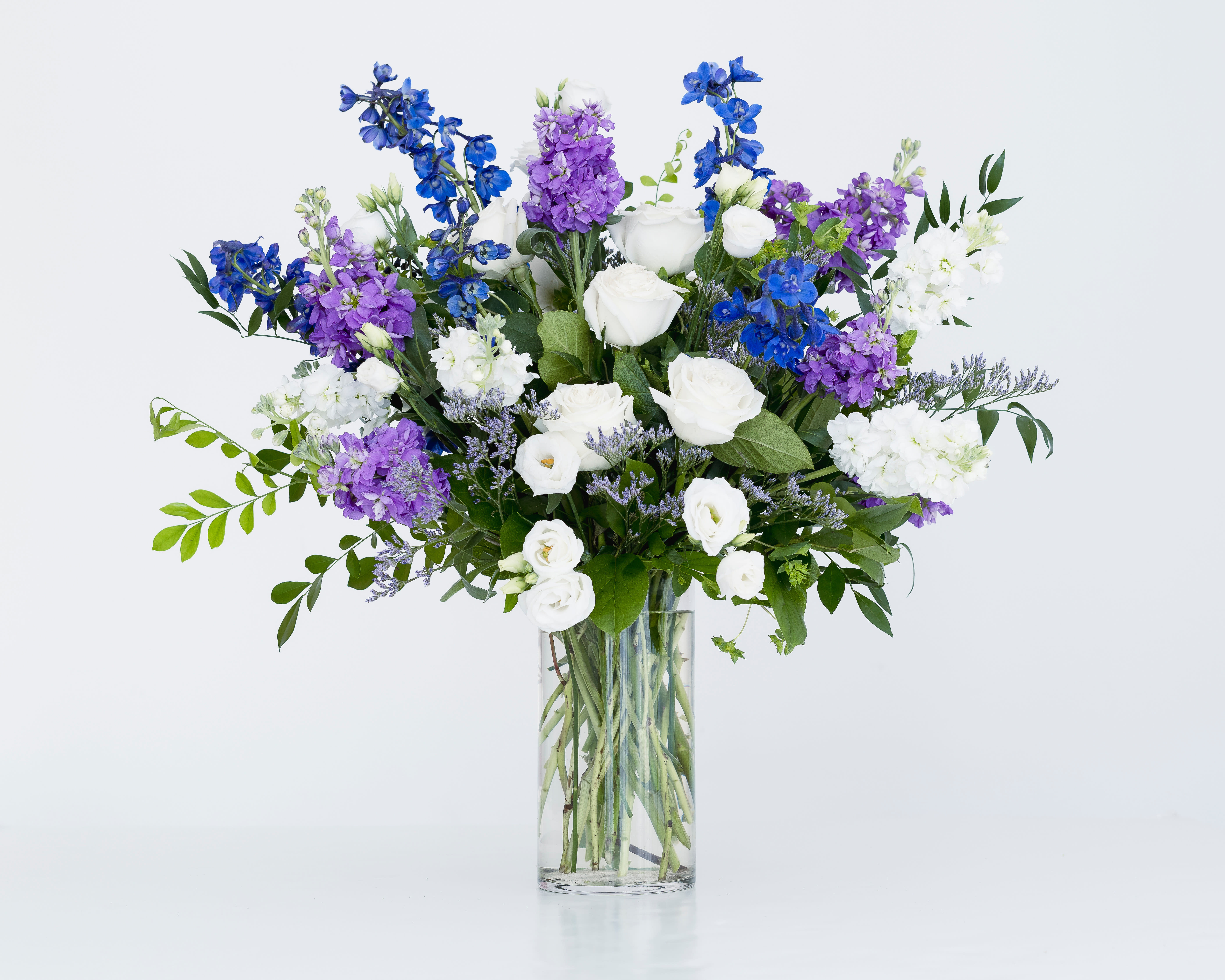 Blue Skies - Signature Size - A gorgeous and dramatic arrangement in shades of blue, purple and white - perfect for that anniversary, birthday or important occasion. The largest design in our Blue Skies Collection - this 24" tall presentation is packed full of roses, Stock, Delphinium and Lisianthus and accents of Statice and fresh greenery all designed in a sleek glass vase. The photo does not do it justice! This piece will send messages of love, joy and thanks in a BIG way!   