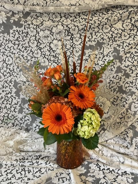 Orange gerbera daisies and roses in a brown vase with a lace backdrop