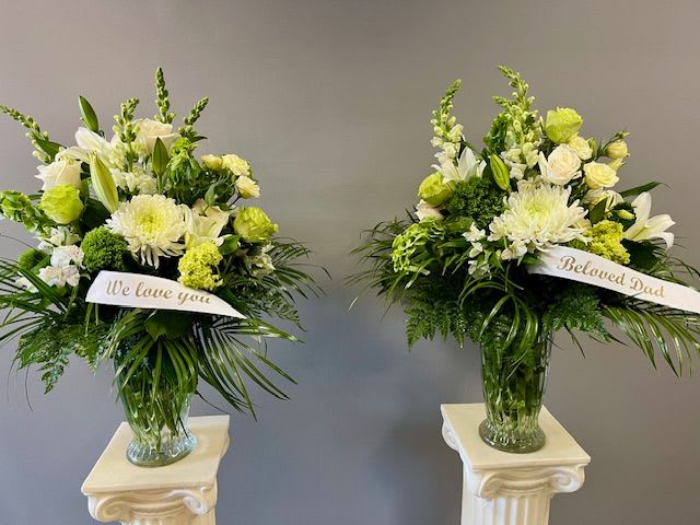 Two white and green sympathy flower arrangements in glass vases with ribbon banners