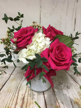 Small arrangement of hot pink roses and white hydrangea in a white textured vase with greenery.