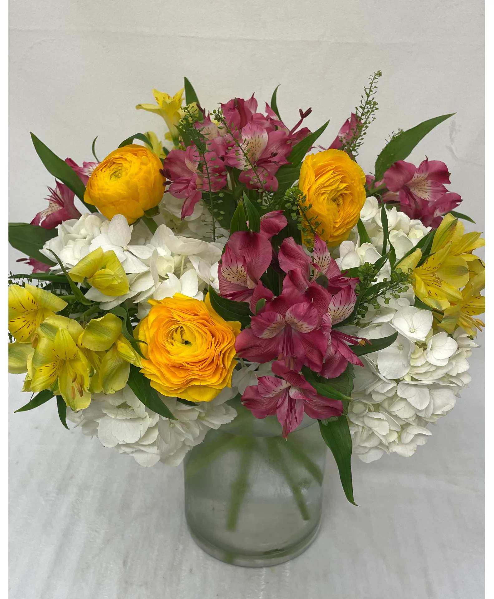 Mixed bouquet of white hydrangeas, yellow blooms, and pink alstroemeria in a clear glass vase