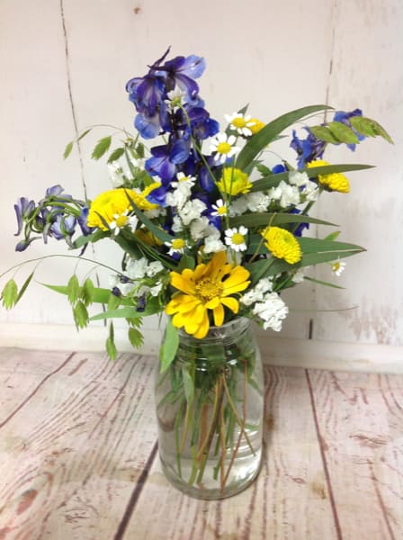Wildflower-style bouquet of blue delphinium, yellow daisies, and white blooms in a clear glass jar vase