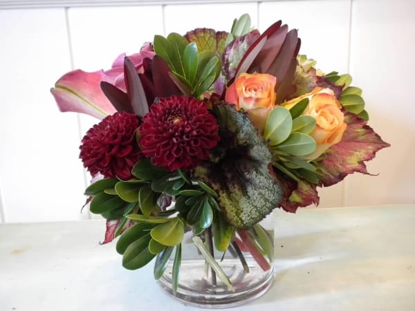 Compact arrangement of burgundy dahlias, peach roses, pink calla lilies, and variegated foliage in a glass vase