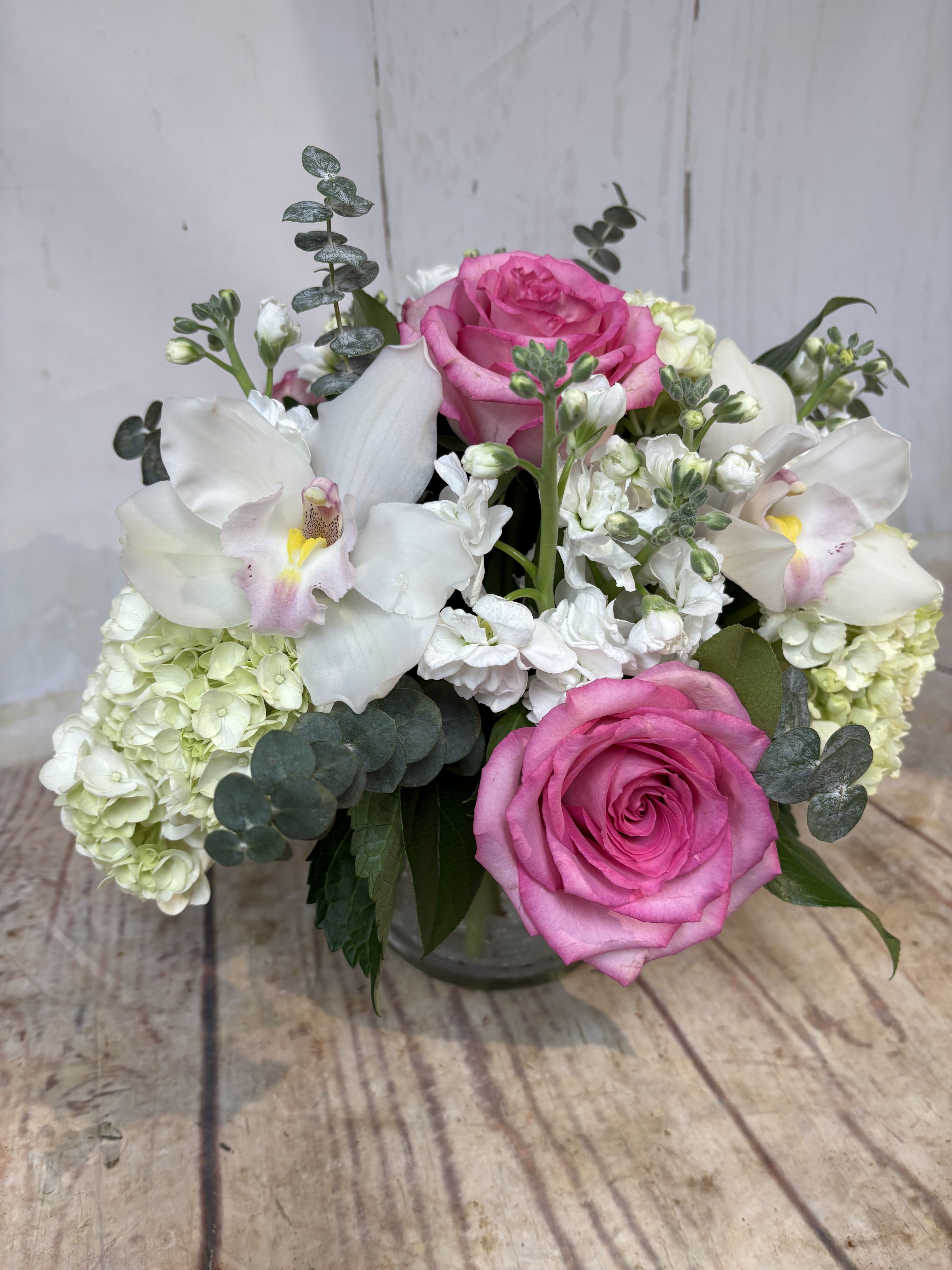 Low arrangement of pink roses, white hydrangeas, stock, and orchids in a clear glass vase
