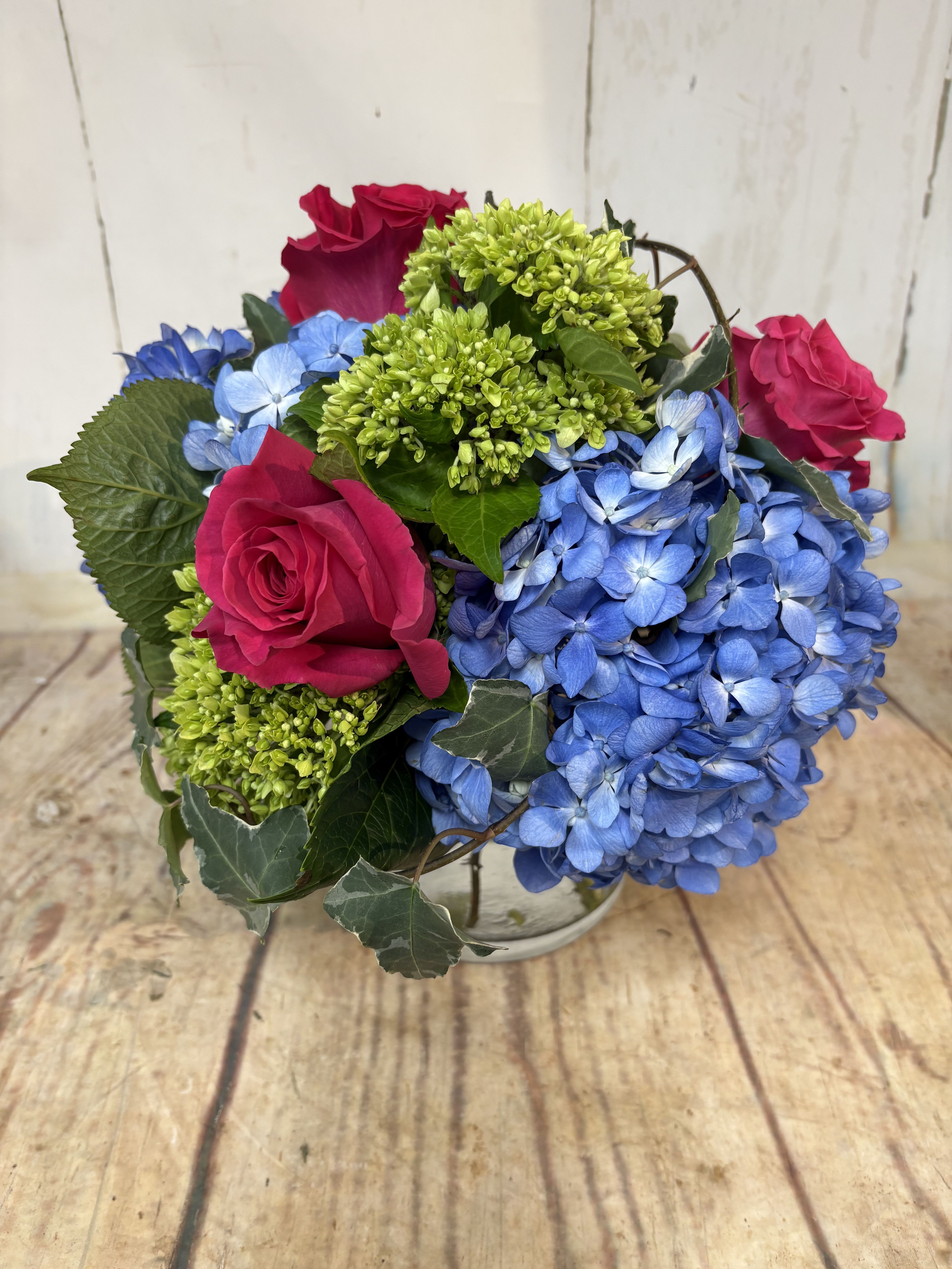 Low round arrangement of hot pink roses, green blooms, and blue hydrangeas in a clear glass vase