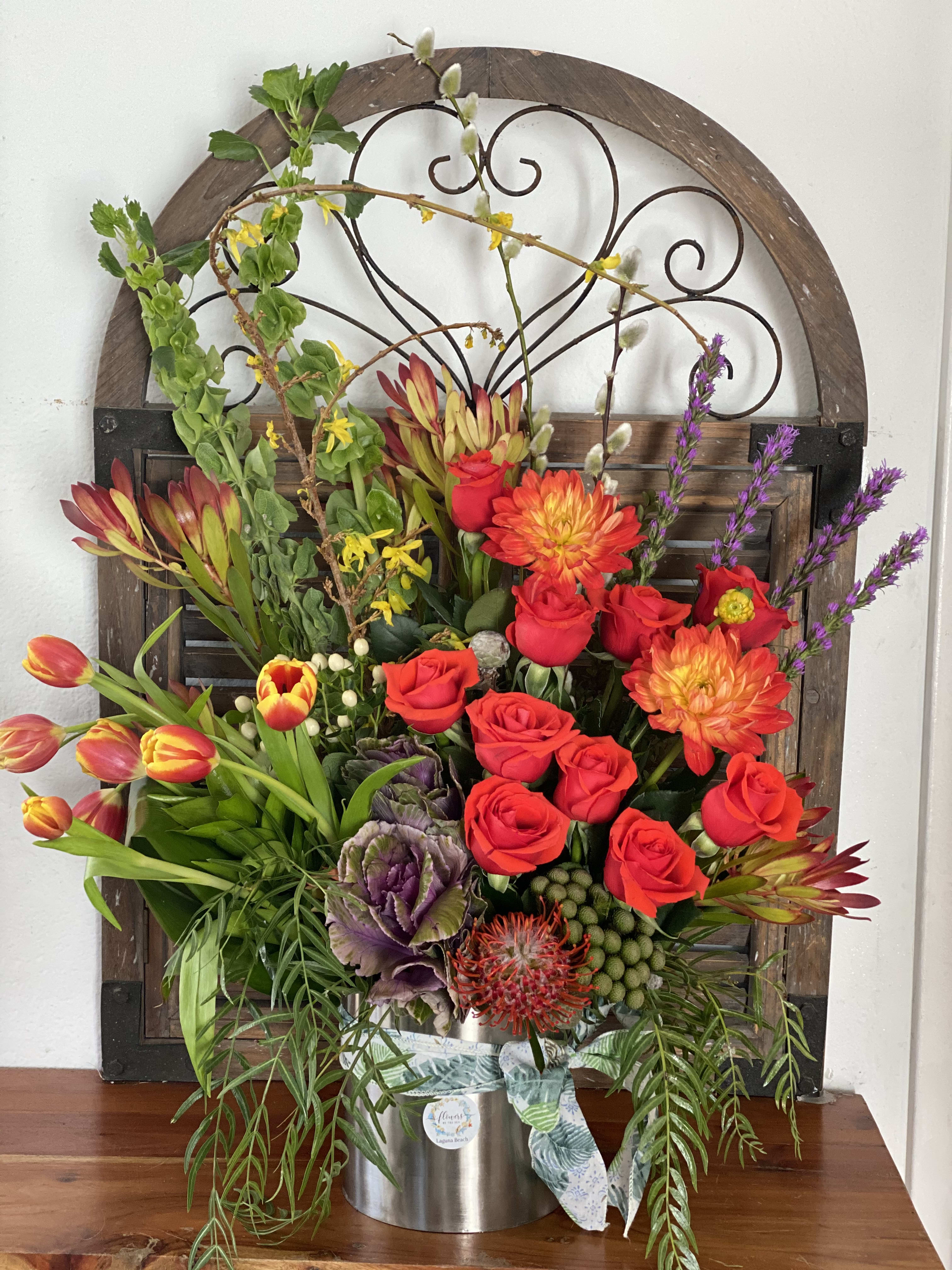 Mixed floral arrangement with red roses, orange dahlias, and tulips in a metal container