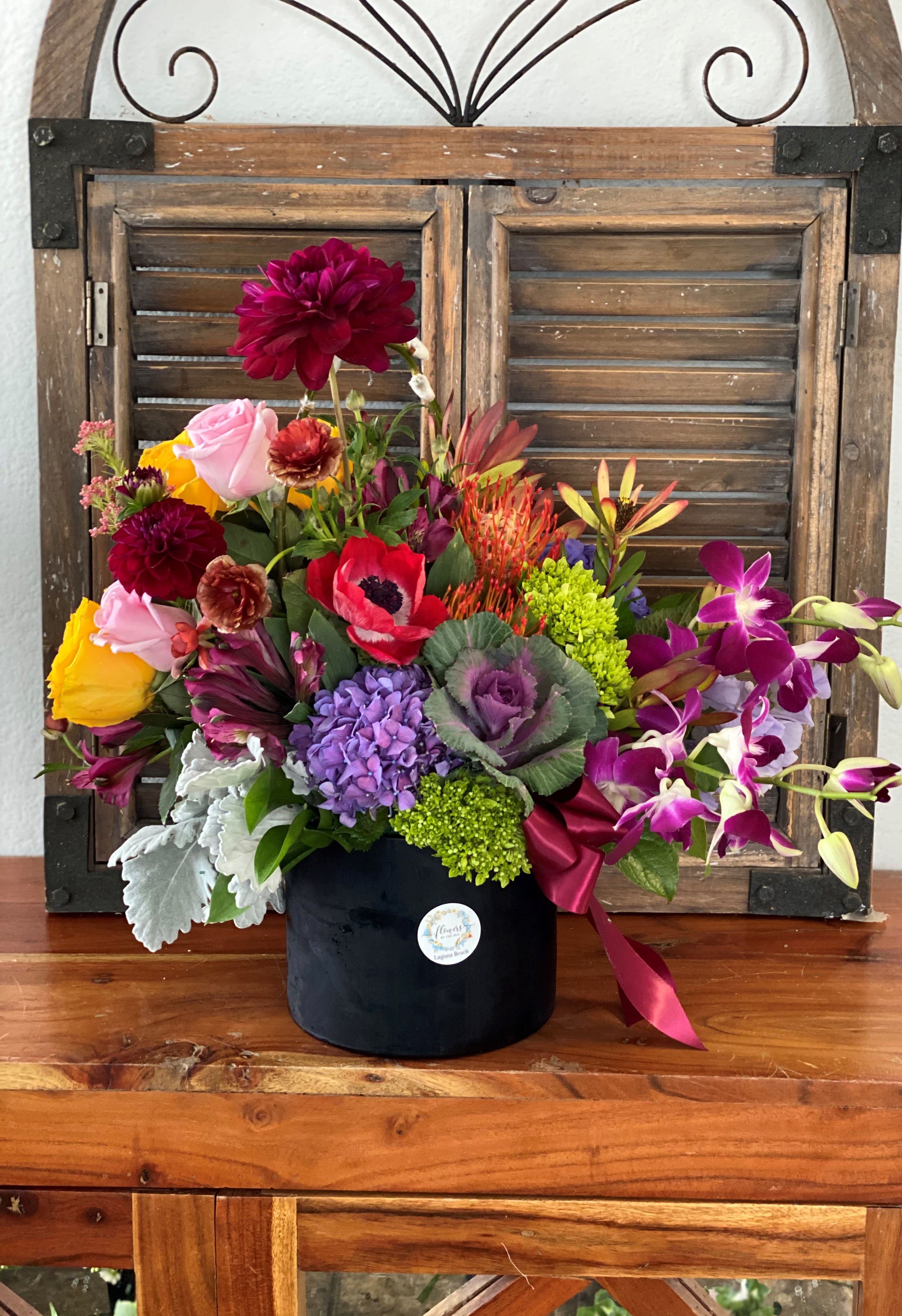 Colorful mixed flower arrangement with roses, orchids, hydrangea, and cabbage in a black cylinder vase
