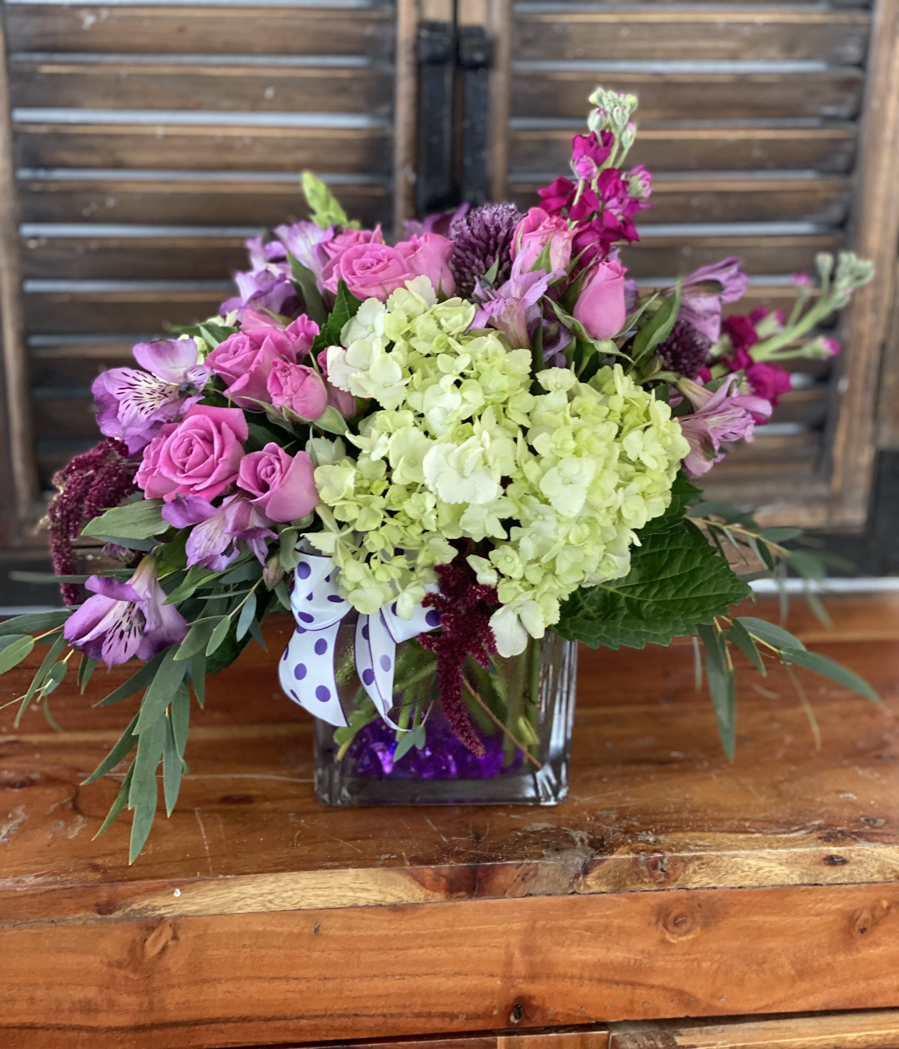 Pink roses and purple flowers in a square glass vase with a polka-dot ribbon.